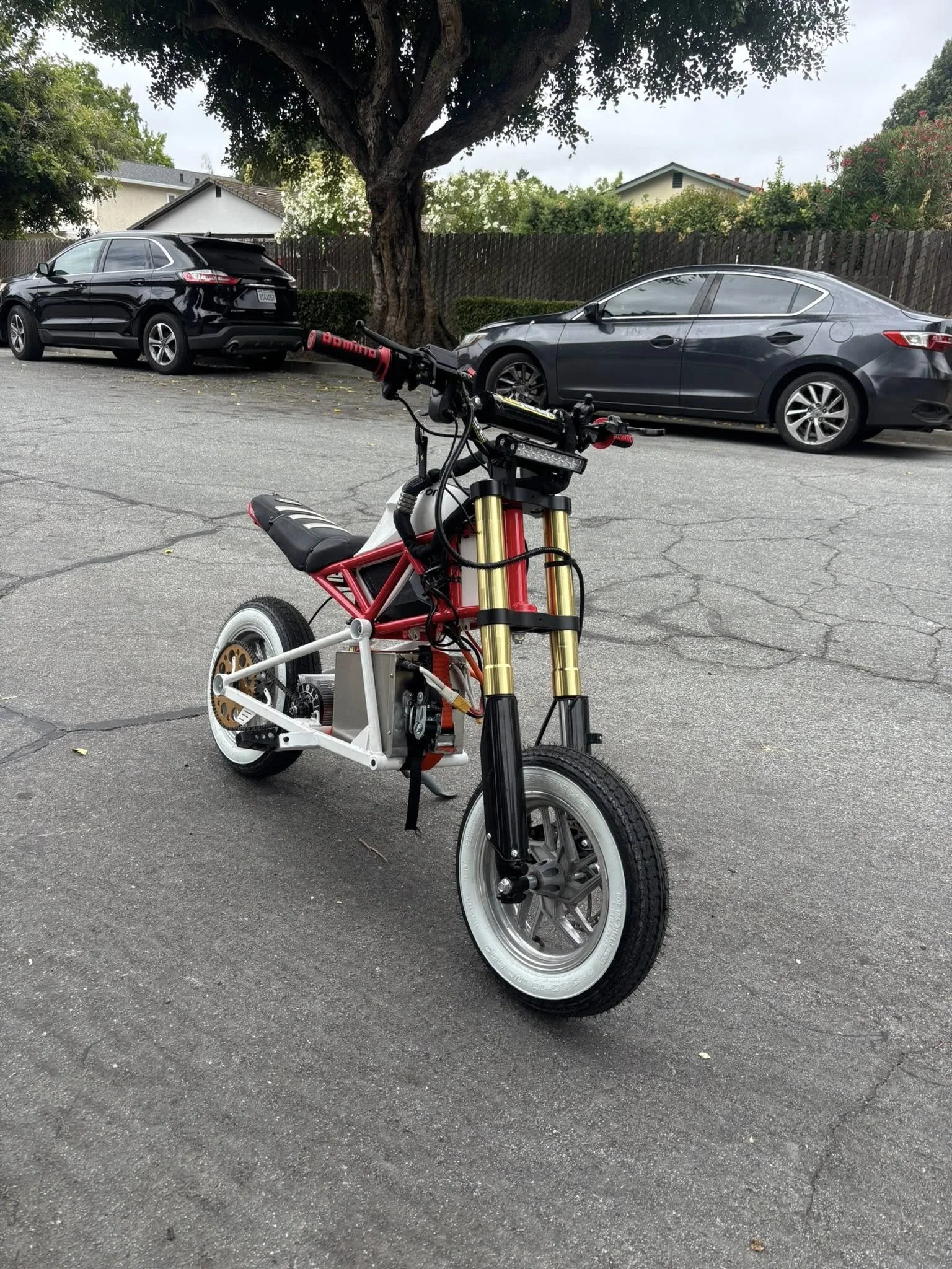 A partially assembled electric bicycle with a black and white frame, black seat, gold front suspension forks, and white tires, parked on an asphalt surface with cars and trees in the background.