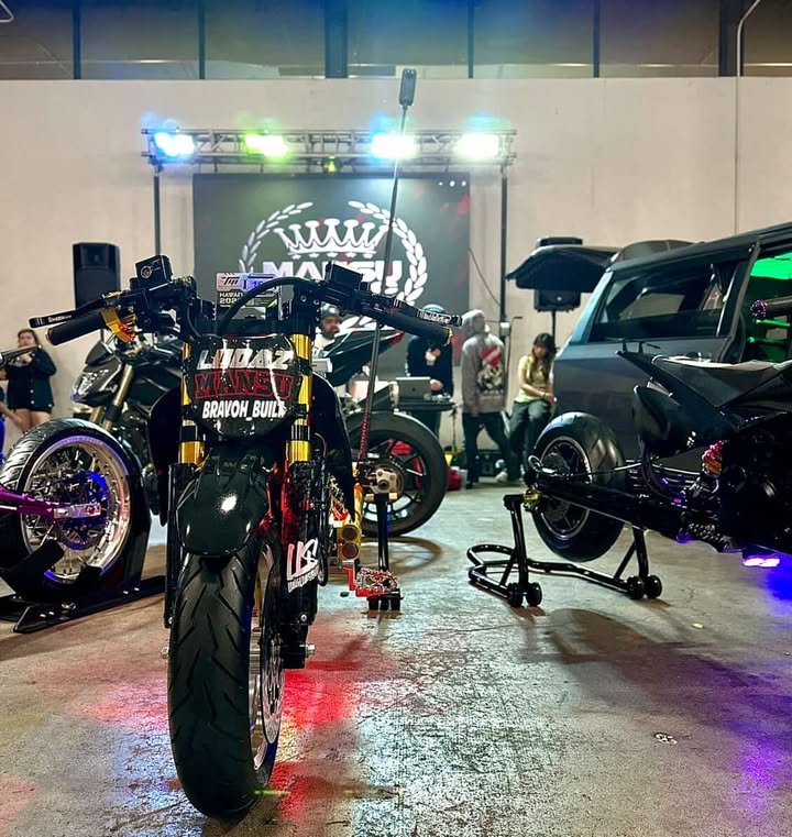 A motorcycle on a platform with a catch and a black car with a wide rear wing at an indoor car show, with people in the background and a large LED screen displaying a logo.