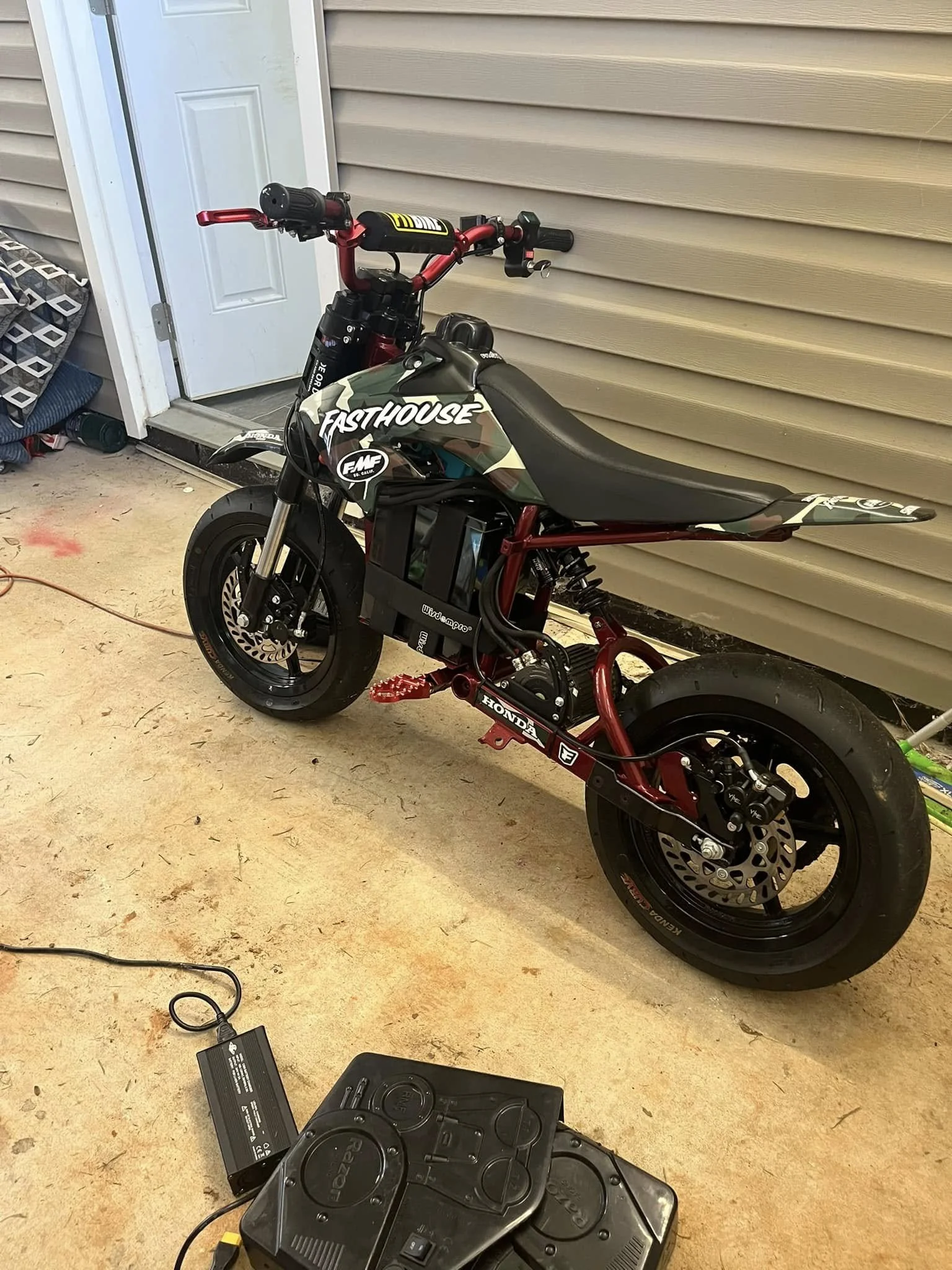 An electric mini motorcycle with a black seat, red frame, and camouflage decals, parked on a garage floor. The motorcycle has disc brakes on both wheels and a black battery pack mounted under the seat. There's a power supply or charger plugged into t