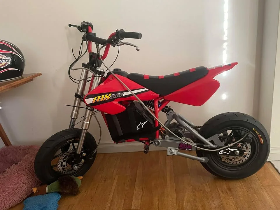 A red MX-500 electric dirt bike with black seat and large black tires, parked indoors against a wall, with a helmet on a nearby shelf.