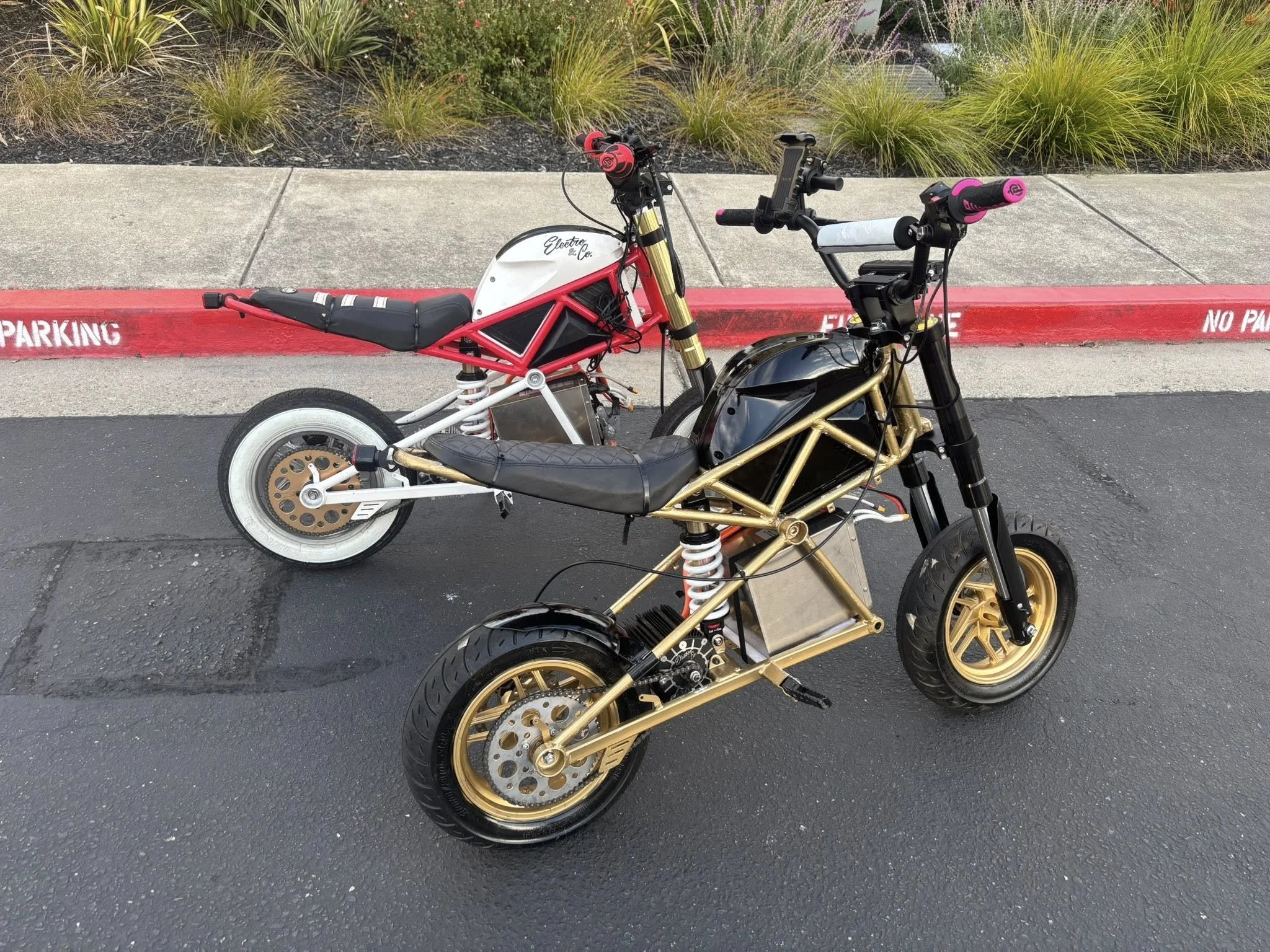 Two electric motorcycles parked on a street near a parking area with bushes and a sidewalk. The motorcycle in the foreground has a gold frame, black seat, and black front tire. The motorcycle in the background has a white frame, black seat, and white