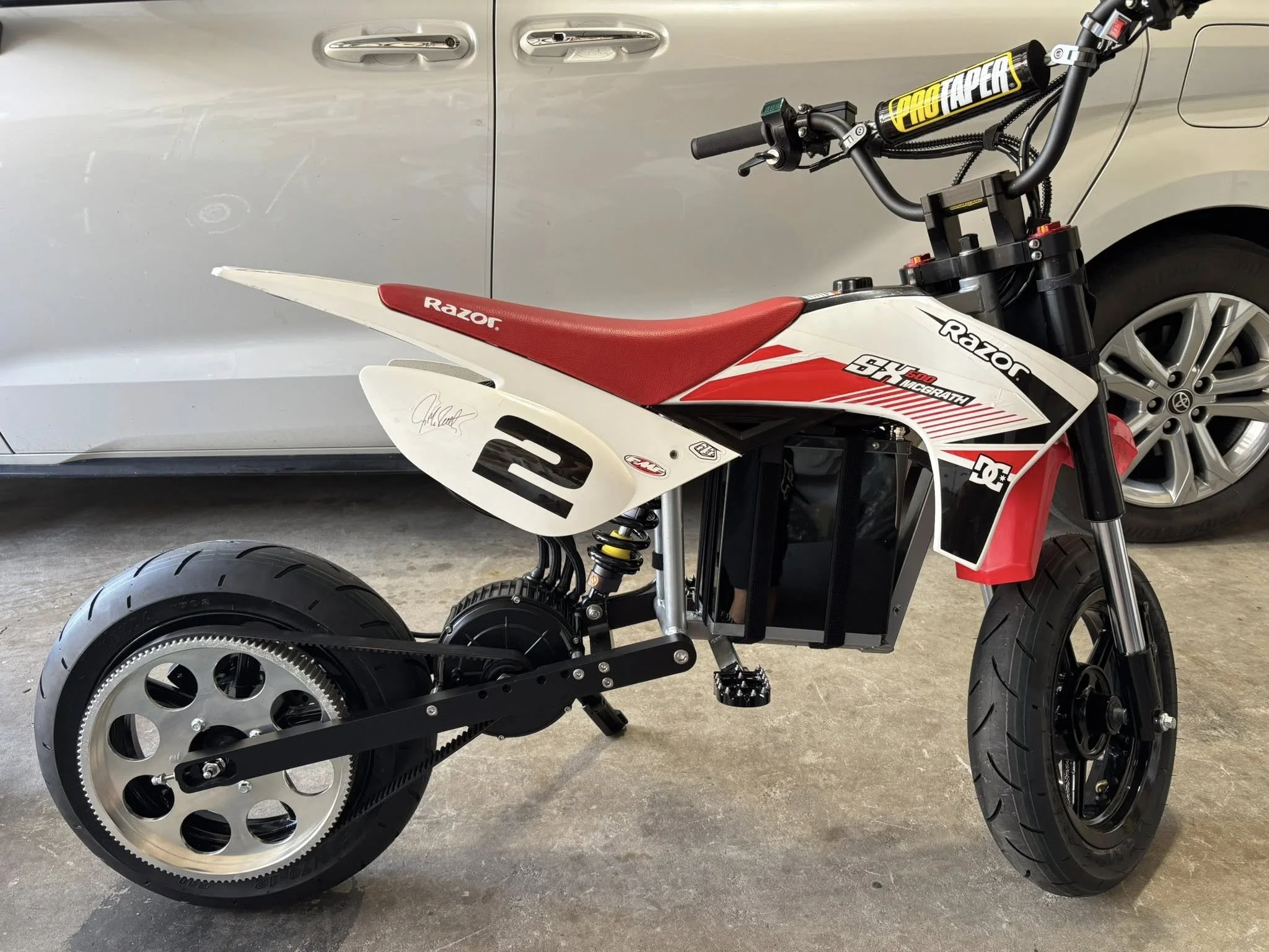 Mini electric motorcycle with a red seat, black handlebars, and a black battery pack, branded with Razor and featuring a DC logo, parked on a concrete garage floor next to a silver car.