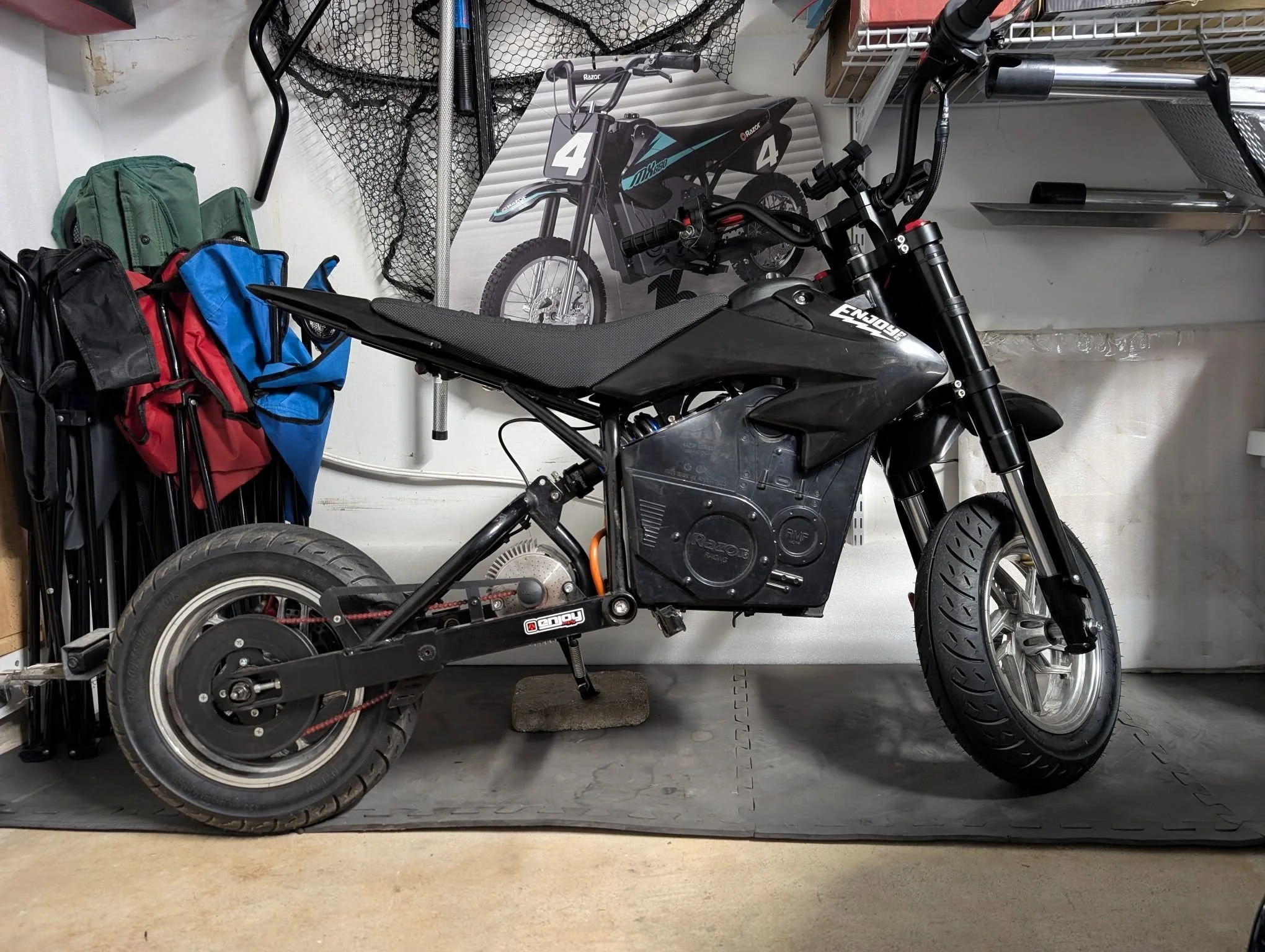 A black electric mini motorcycle with a black seat, set on a garage floor. In the background, there are red, blue, and green backpacks, a poster of a dirt bike, and some hanging and shelving storage.