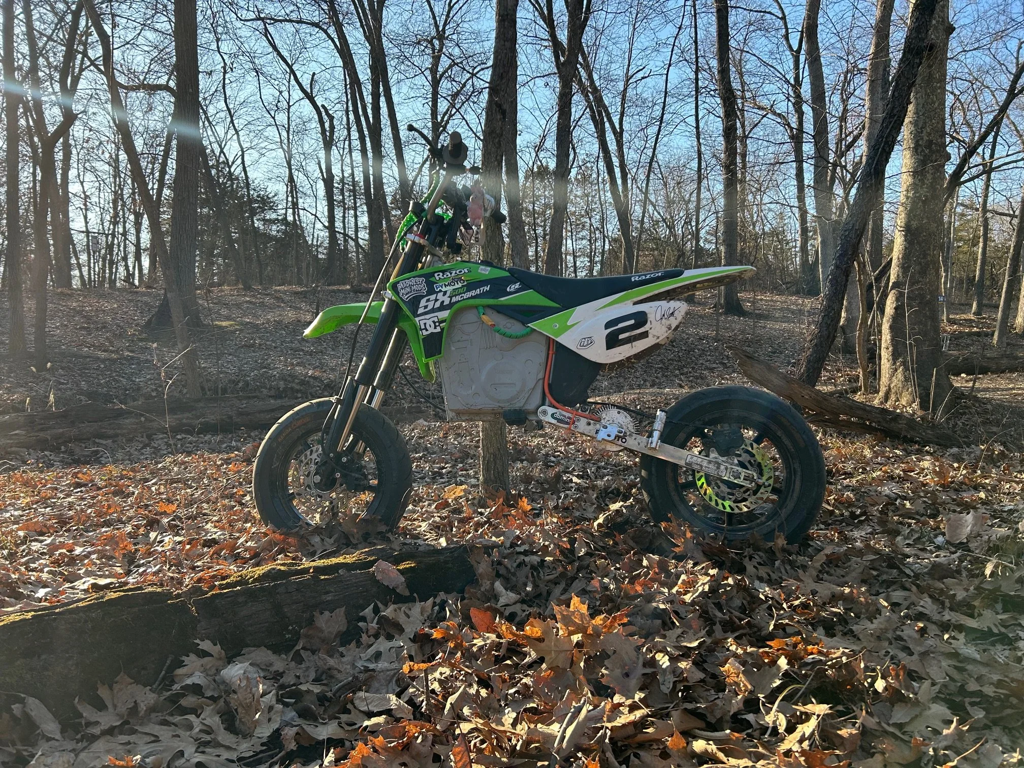 A green and black dirt bike with the number 2 on its side, sitting on a forest floor covered in fallen leaves.