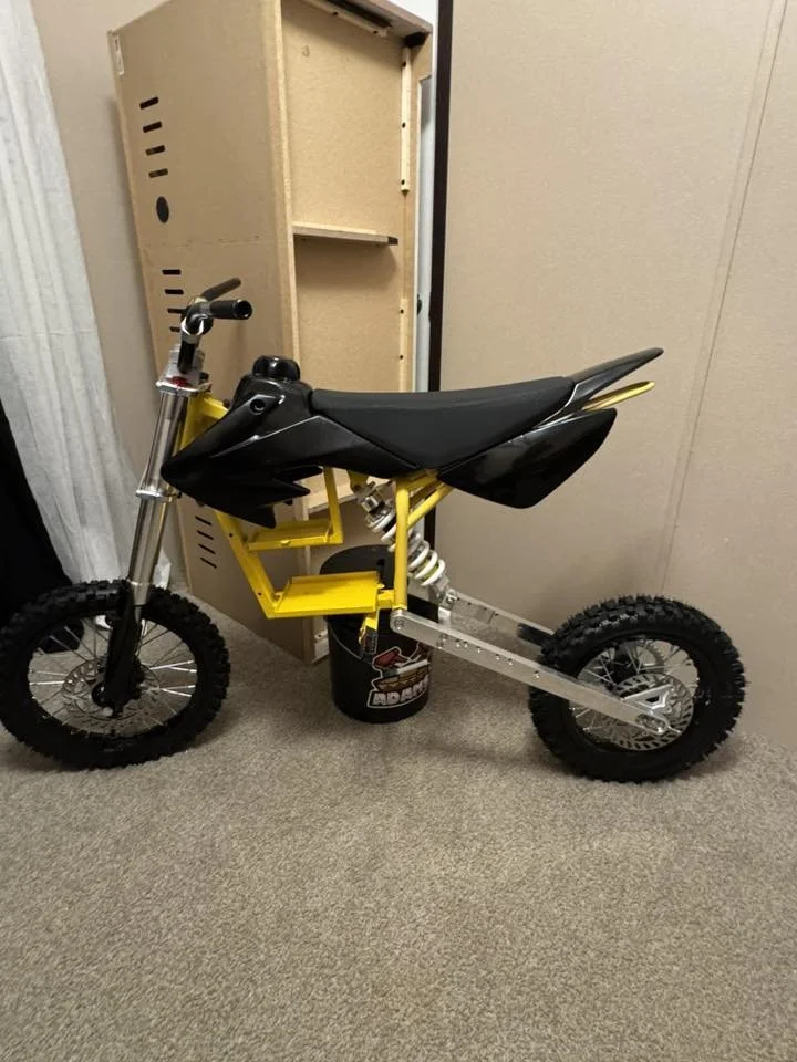 A small dirt bike with a yellow frame, black seat, and off-road tires, positioned indoors next to a beige wall and a wooden storage shelf.