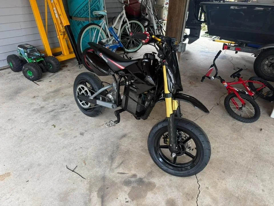 A partially assembled black electric motorcycle with yellow front forks, parked on a concrete floor in a garage with three children's bikes, a toy car, and a pickup truck in the background.