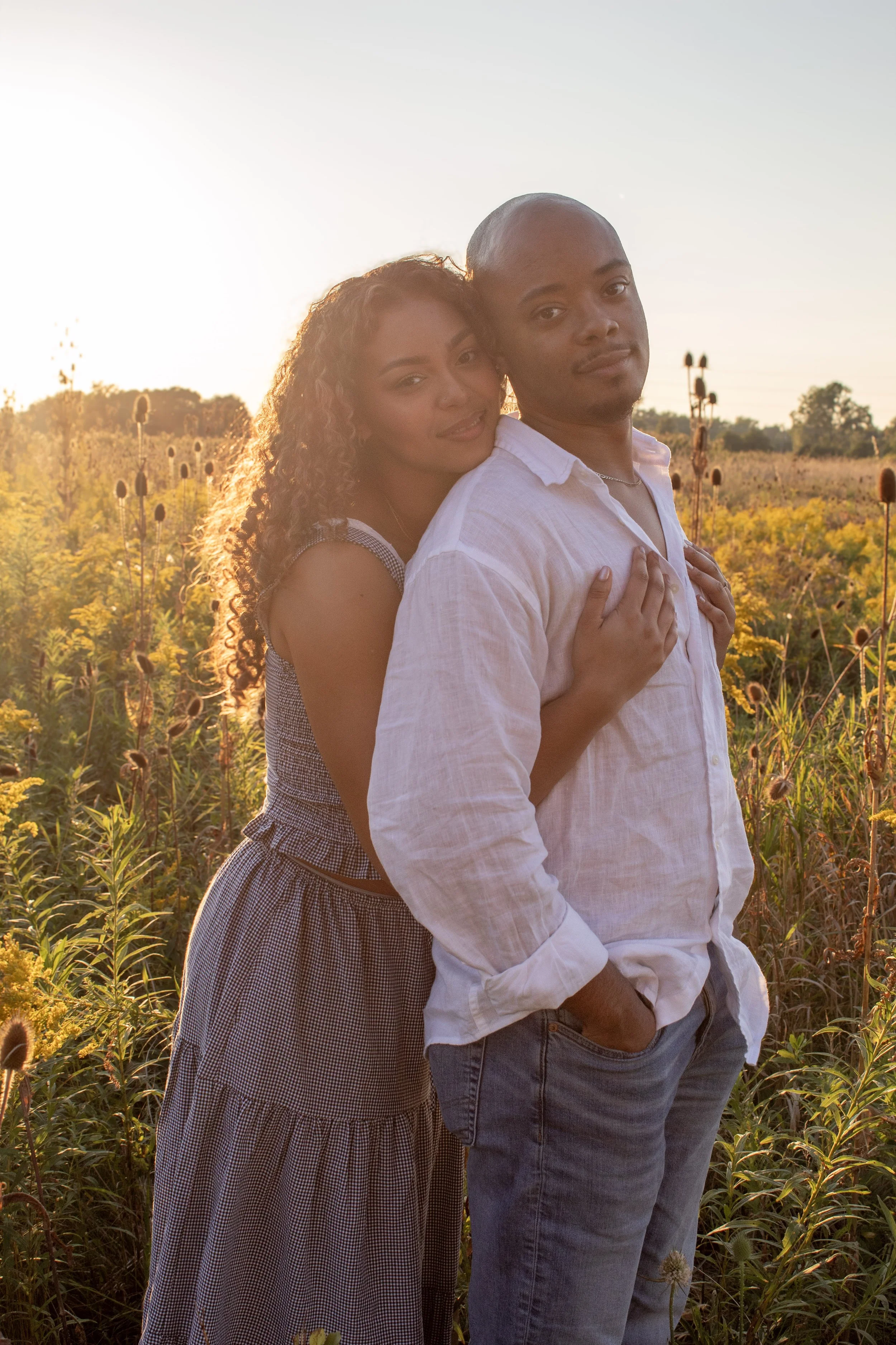 A young couple standing close together in a field of yellow flowers at sunset. The woman is hugging the man from behind, with her arms around his shoulders, and both are smiling softly. The man is wearing a white shirt, and the woman is in a checkere