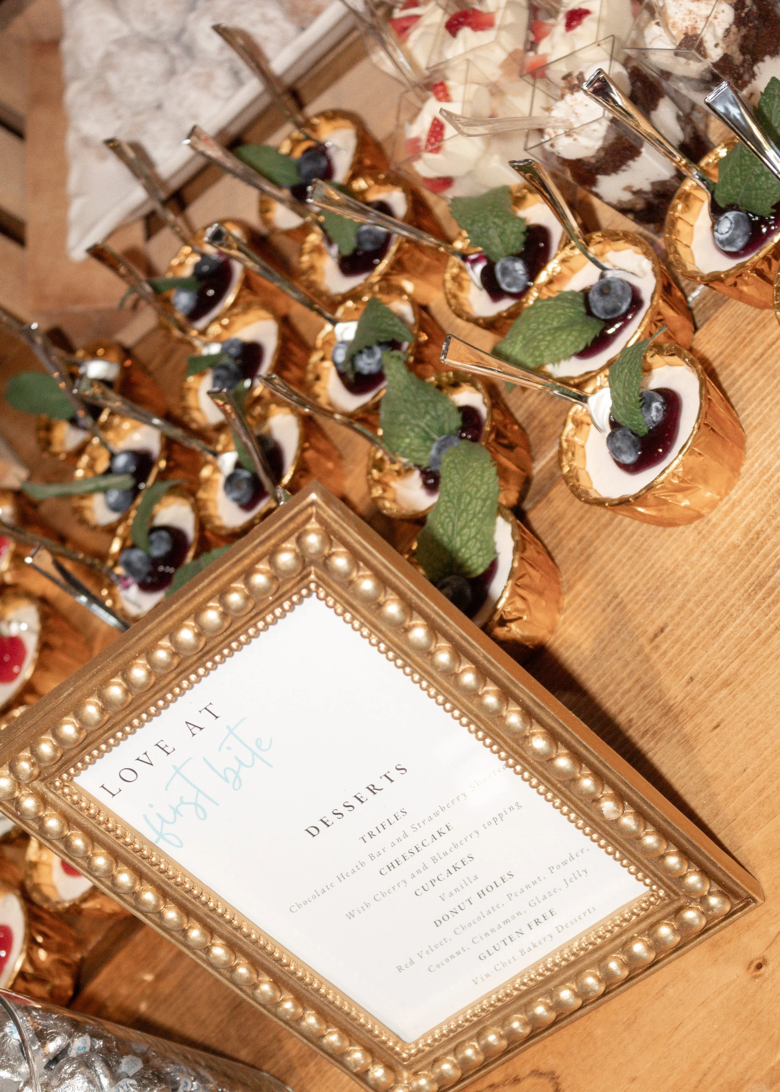 A framed dessert menu on a wooden table, with mini cheesecakes topped with blueberries and mint leaves, and other desserts visible in the background.