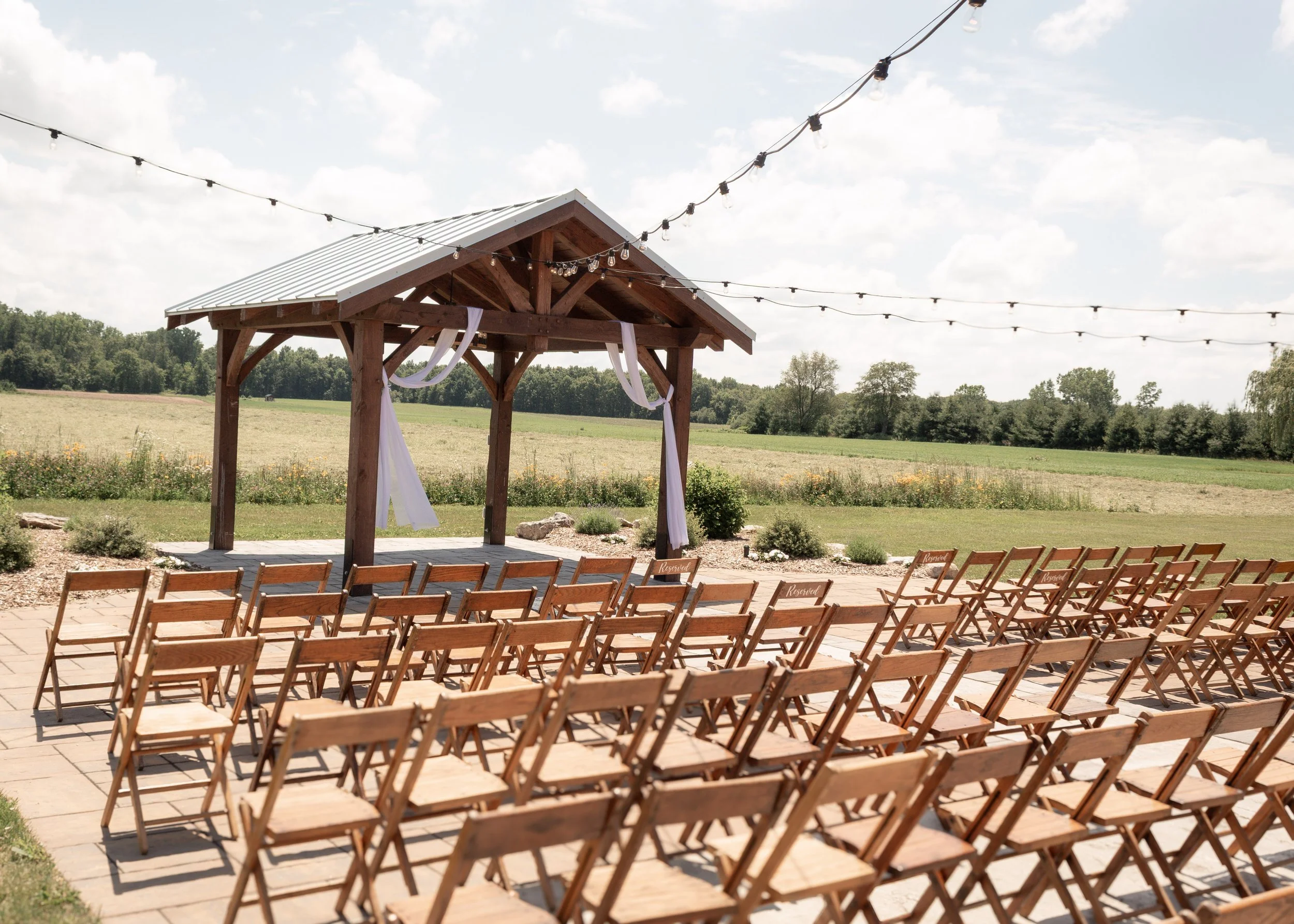 Outdoor wedding ceremony setup with rows of wooden chairs facing a wooden arch decorated with white fabric, set in a grassy field under a partly cloudy sky with string lights overhead.