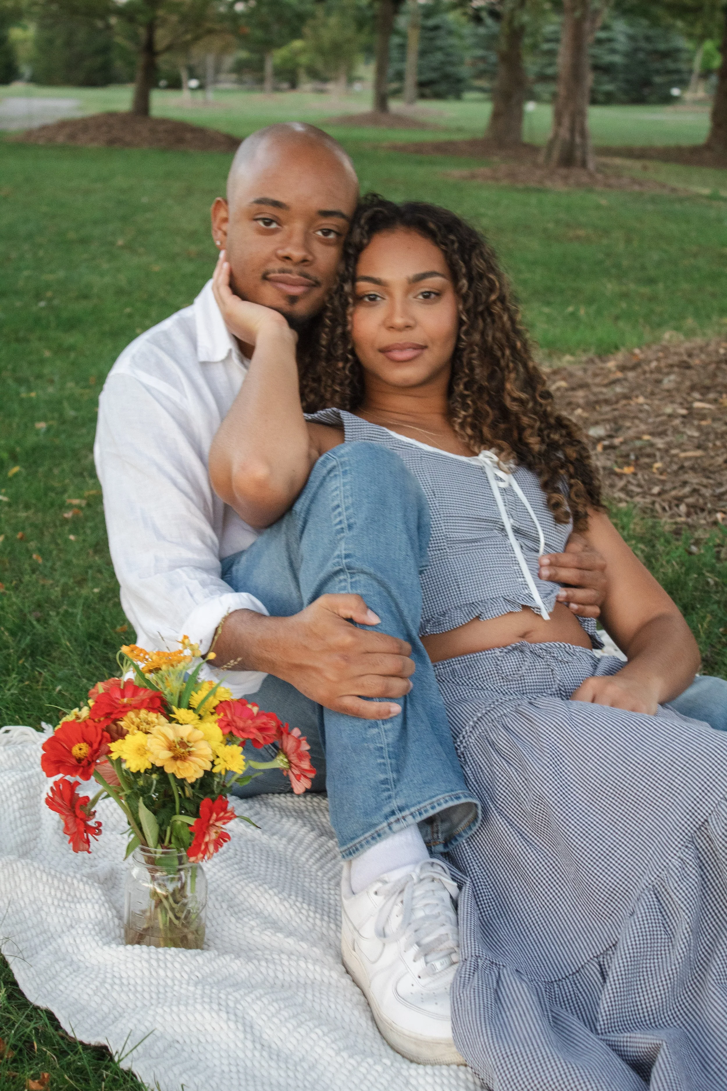 A young couple sitting on a blanket in a park, surrounded by trees. They are posing closely with the man gently holding the woman. The woman has curly hair and is wearing a blue checkered outfit, while the man is bald and is wearing a light-colored s