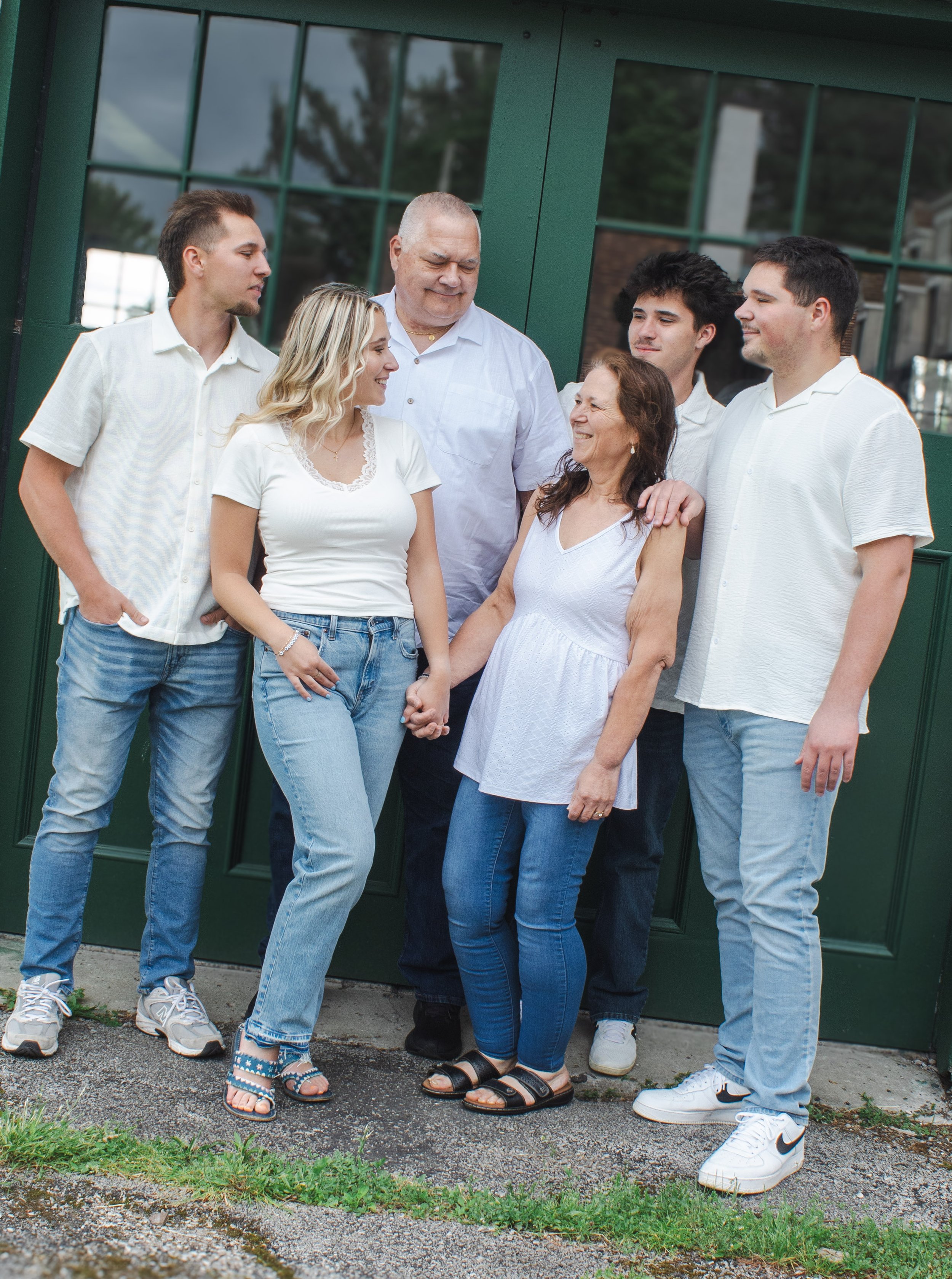 A multigenerational family of six standing together outside near a green door, holding hands and smiling warmly. The group consists of three men, two women, and a teenage girl, all dressed casually.