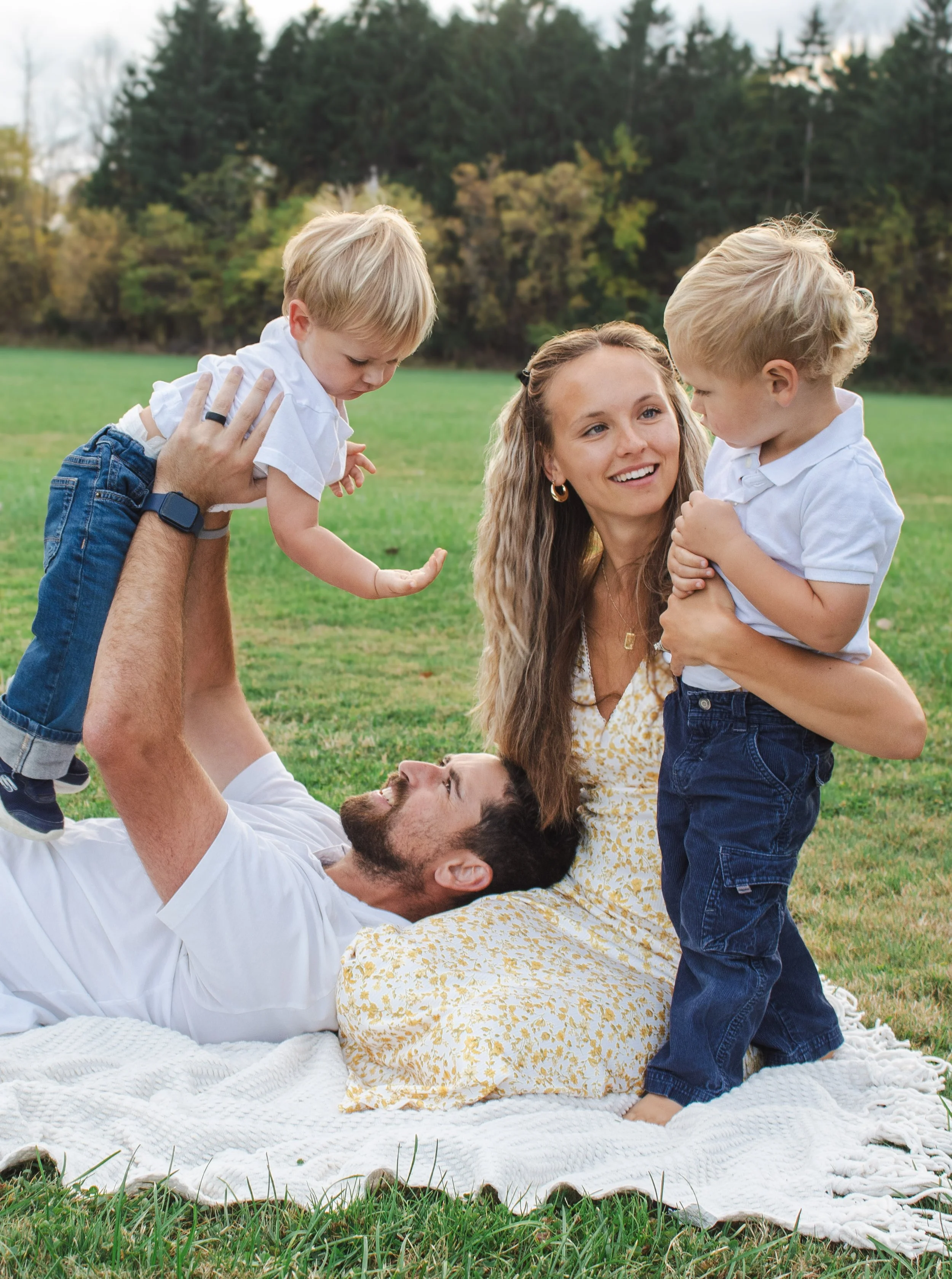 A family of four enjoys a playful moment outdoors on a grassy field, with trees in the background. A man lies on his back on a white blanket, lifting a young boy with blonde hair and a white shirt into the air. The woman, with long brown hair, sits nearby smiling at the boy. A younger boy with blonde hair, also in a white shirt, stands close to her, being held by her. The scene captures a joyful, familial interaction in a natural setting.