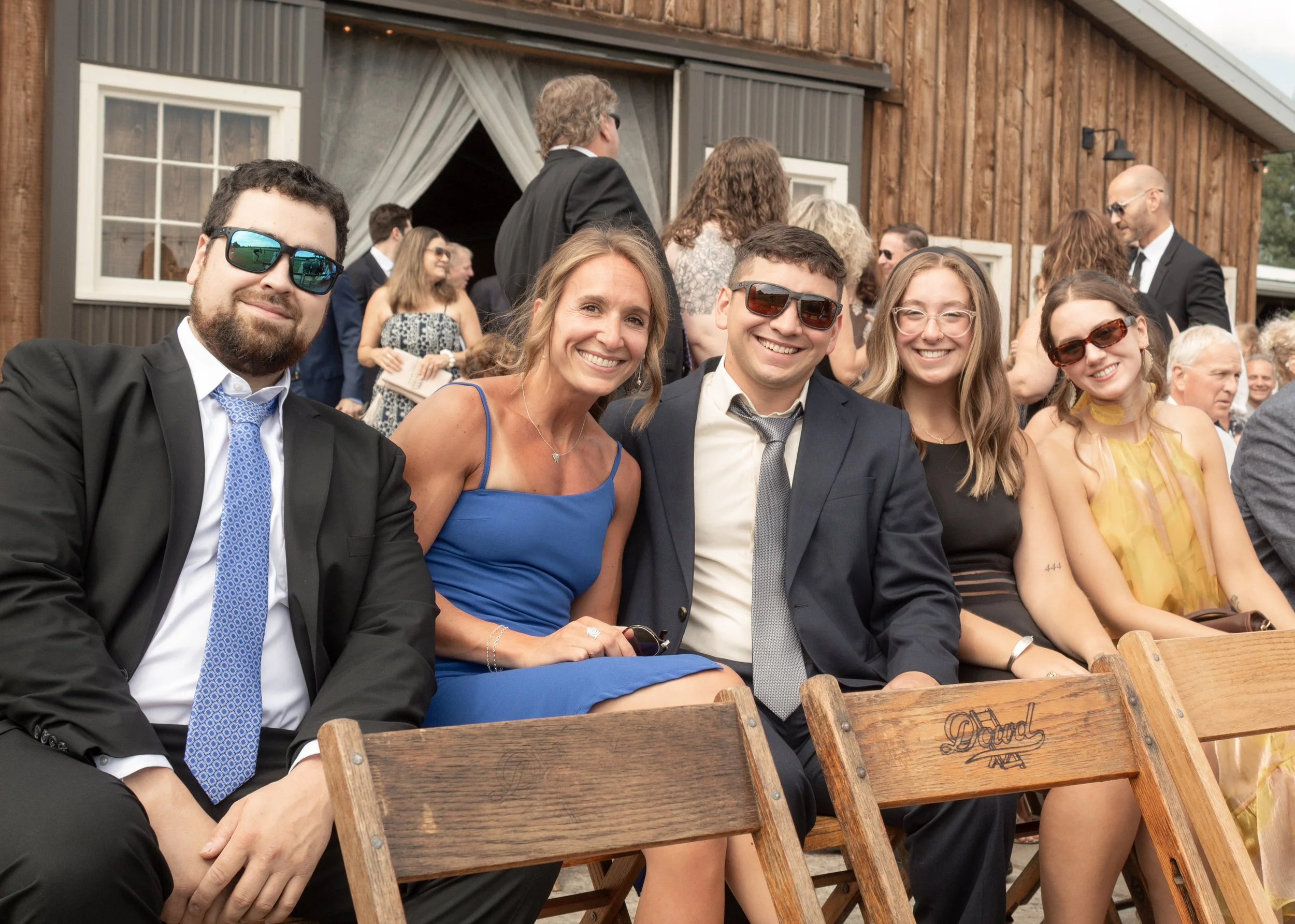Group of people at an outdoor social event, sitting and standing near a wooden building, dressed in formal and semi-formal attire, smiling, some wearing sunglasses, with a rustic ambiance.