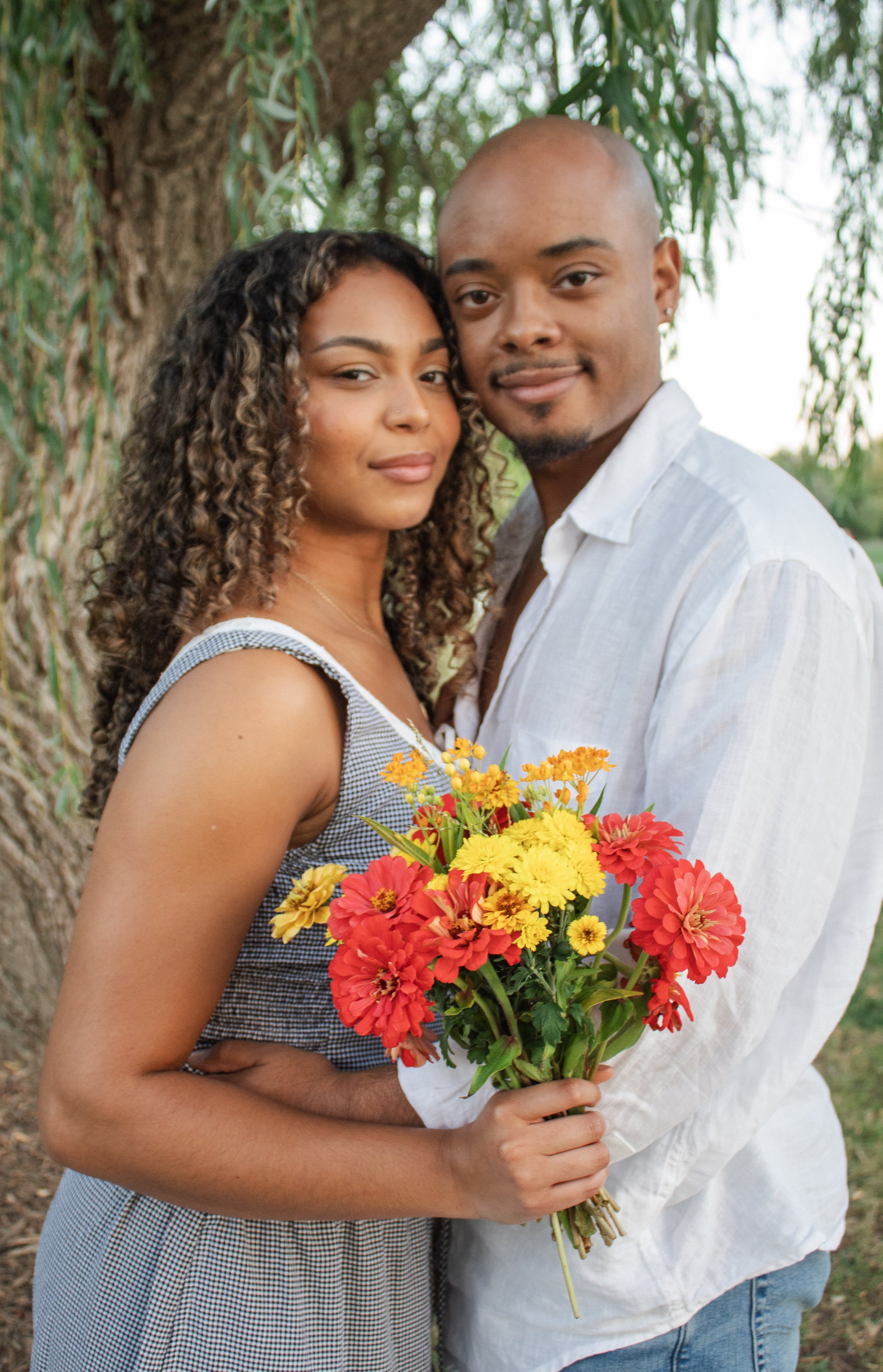 A young couple stands outdoors near a large tree, with the woman holding a vibrant bouquet of yellow, red, and orange flowers, both smiling and embracing.