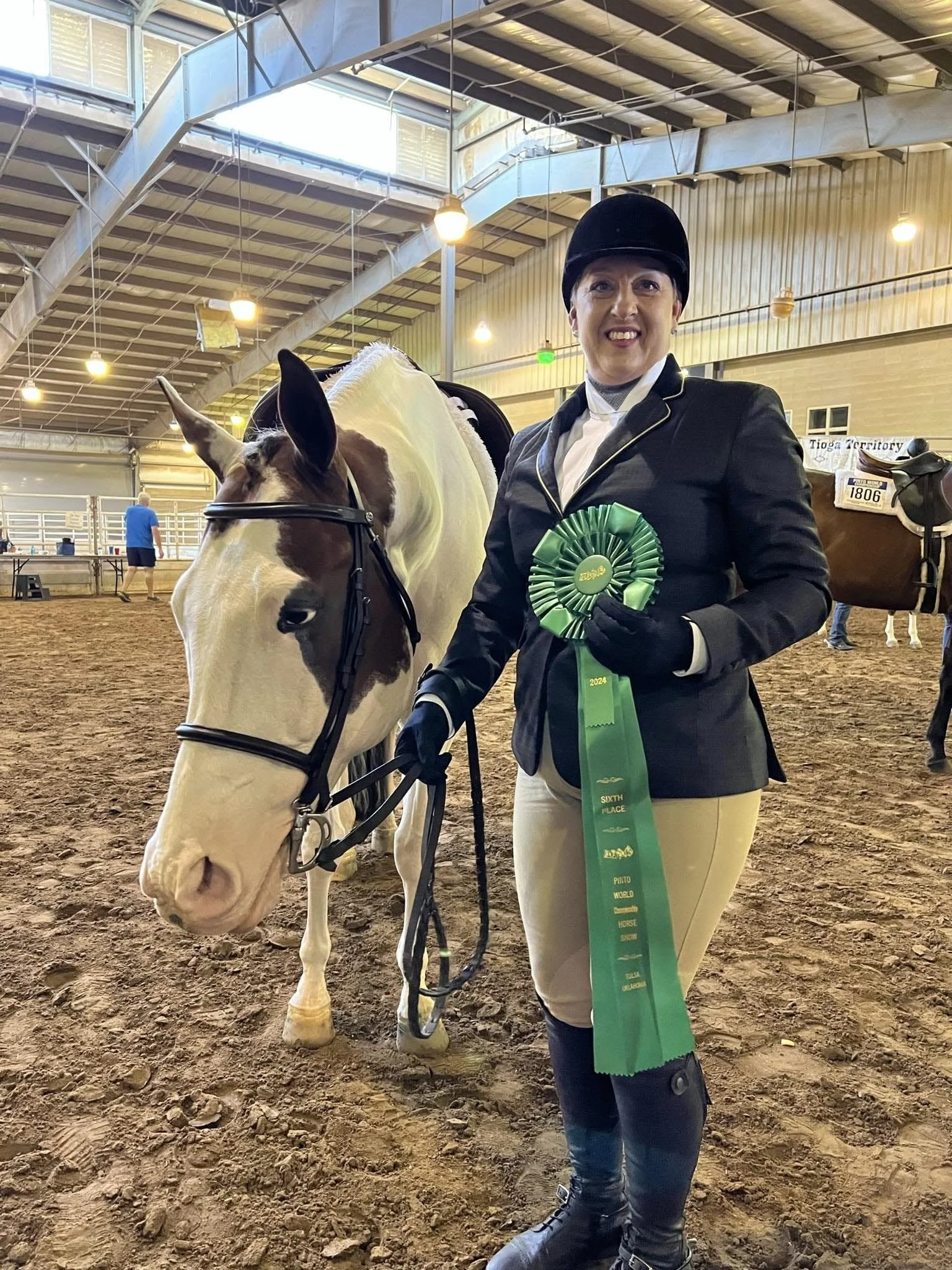 A woman wearing riding attire, including a black helmet, white shirt, black jacket, and beige riding pants, stands inside an indoor riding arena, holding a green ribbon. Next to her is a white horse with brown markings on its face, wearing a bridle. The arena has a dirt floor and is illuminated with overhead lights, with people and additional horses in the background.