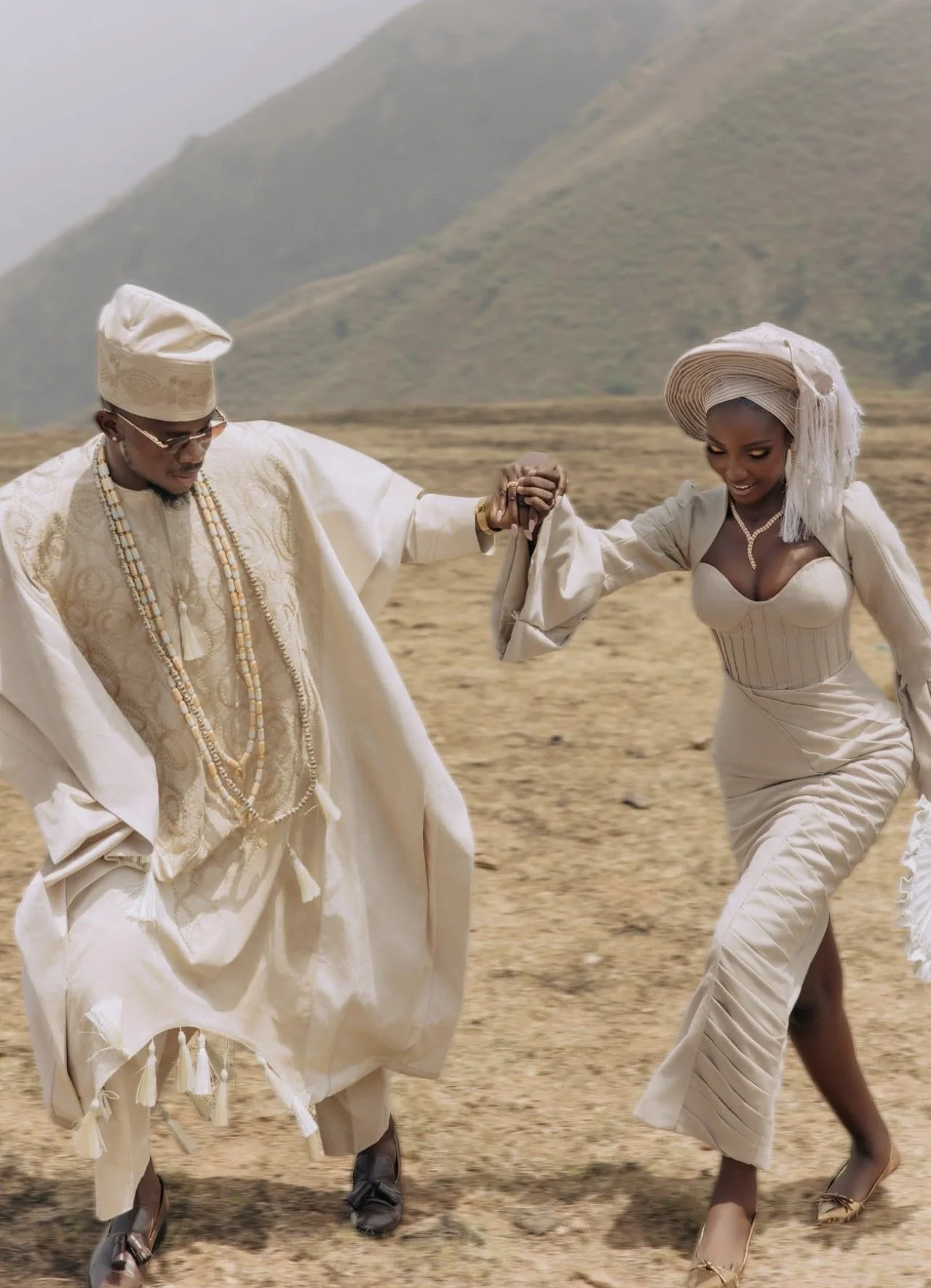 A couple dressed in traditional Nigerian attire, with the woman wearing purple and gold outfit and the man in purple agbada and cap, at a celebration or event.