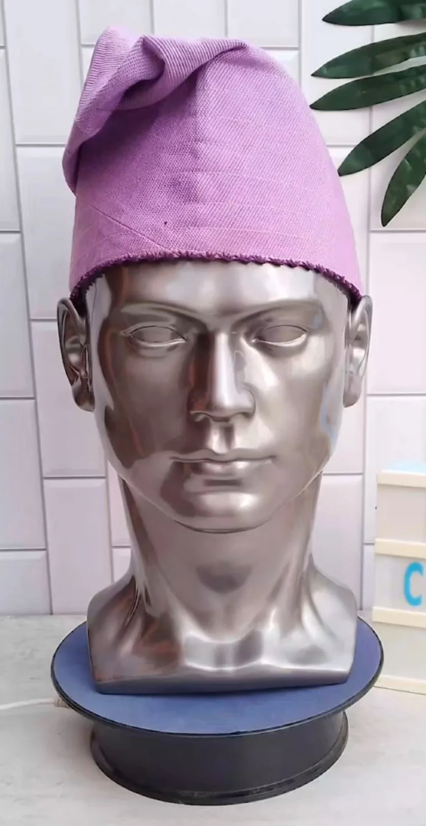 Silver mannequin head wearing a pink knit fila cap with a folded top, placed on a round blue stand, with white tiled wall and green plant in the background.