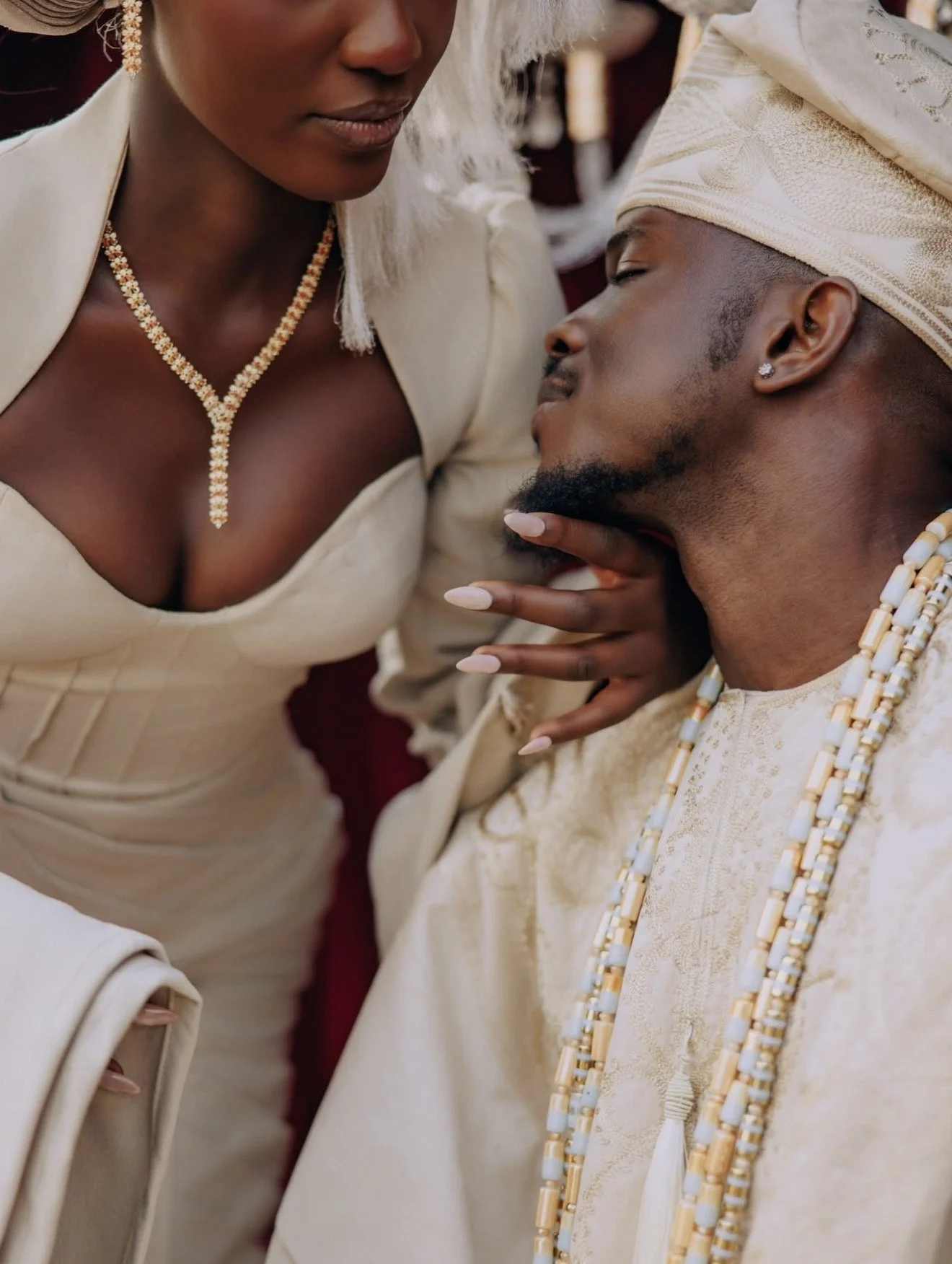 A woman and man dressed in traditional Nigerian attire, standing close together indoors near a glass railing with a city view in the background.