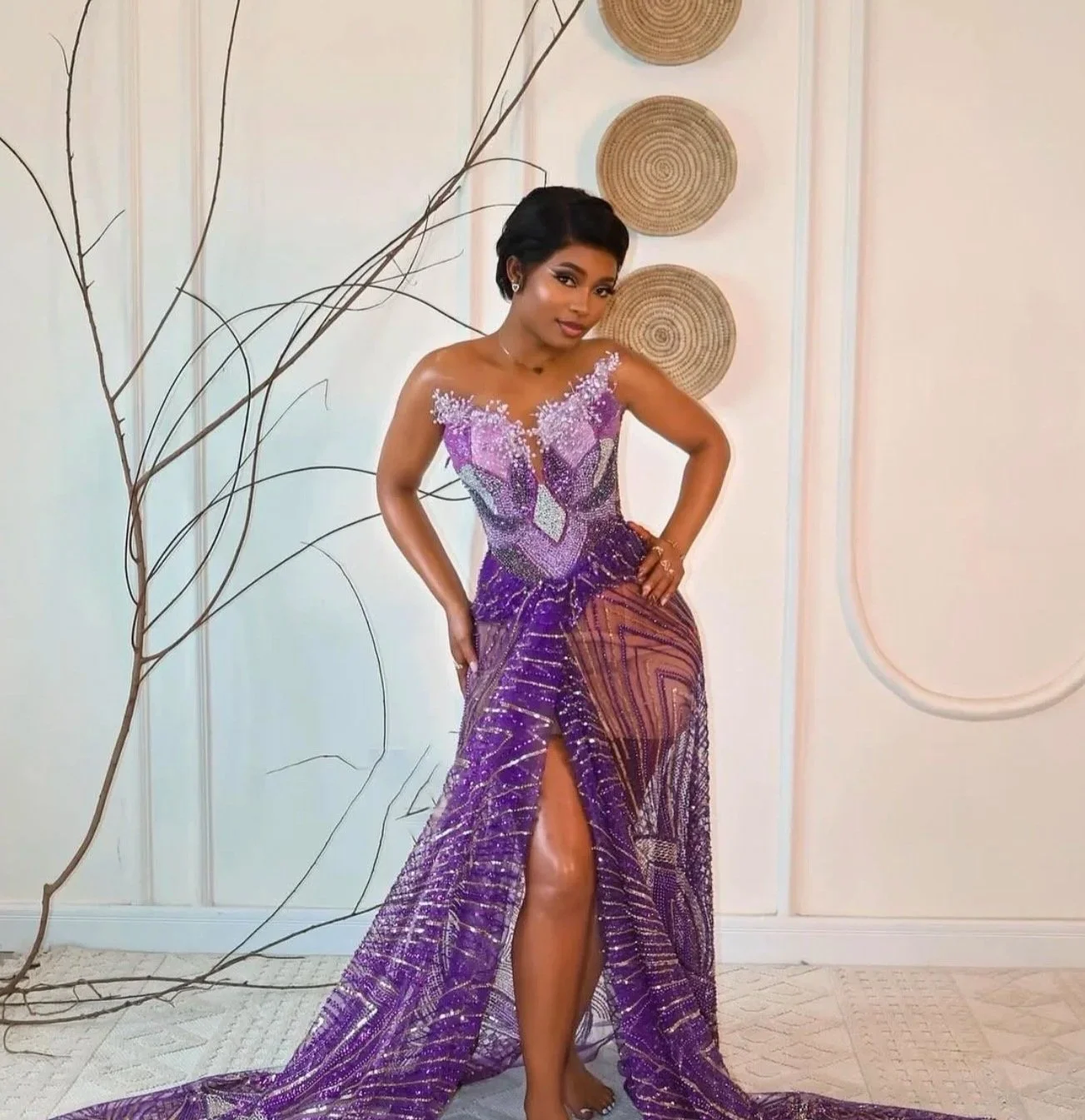 A woman in a purple evening gown with intricate beadwork, a thigh-high slit, and floral detailing, standing indoors against a light-colored wall with decorative wall art and dried plant arrangements.