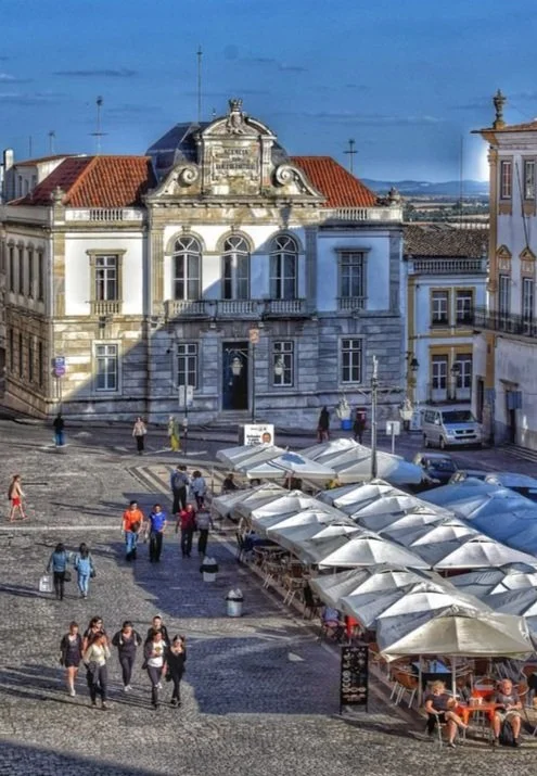 Rua movimentada com pessoas caminhando, vendadores de rua com bancas de pratos e cadeiras sob guarda-sóis brancos, prédio antigo de arquitetura clássica ao fundo, céu azul