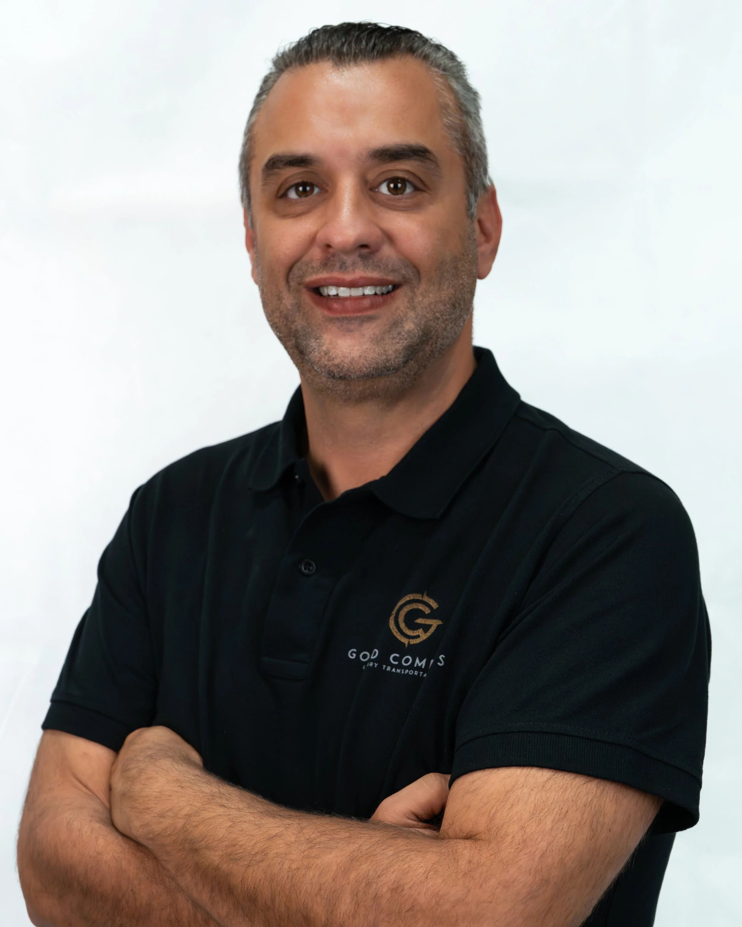 Homem sorridente de cabelos grisalhos, pele clara, usando camiseta preta com logotipo e texto da empresa 'God Comes Cargo Transportes', com braços cruzados, frente a fundo branco.