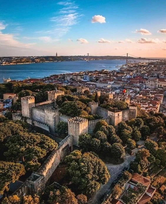 Vista aérea do Castelo de São Jorge em Lisboa, com muros medievais cercando áreas verdes, ao fundo um rio, uma ponte e a cidade ao entardecer.