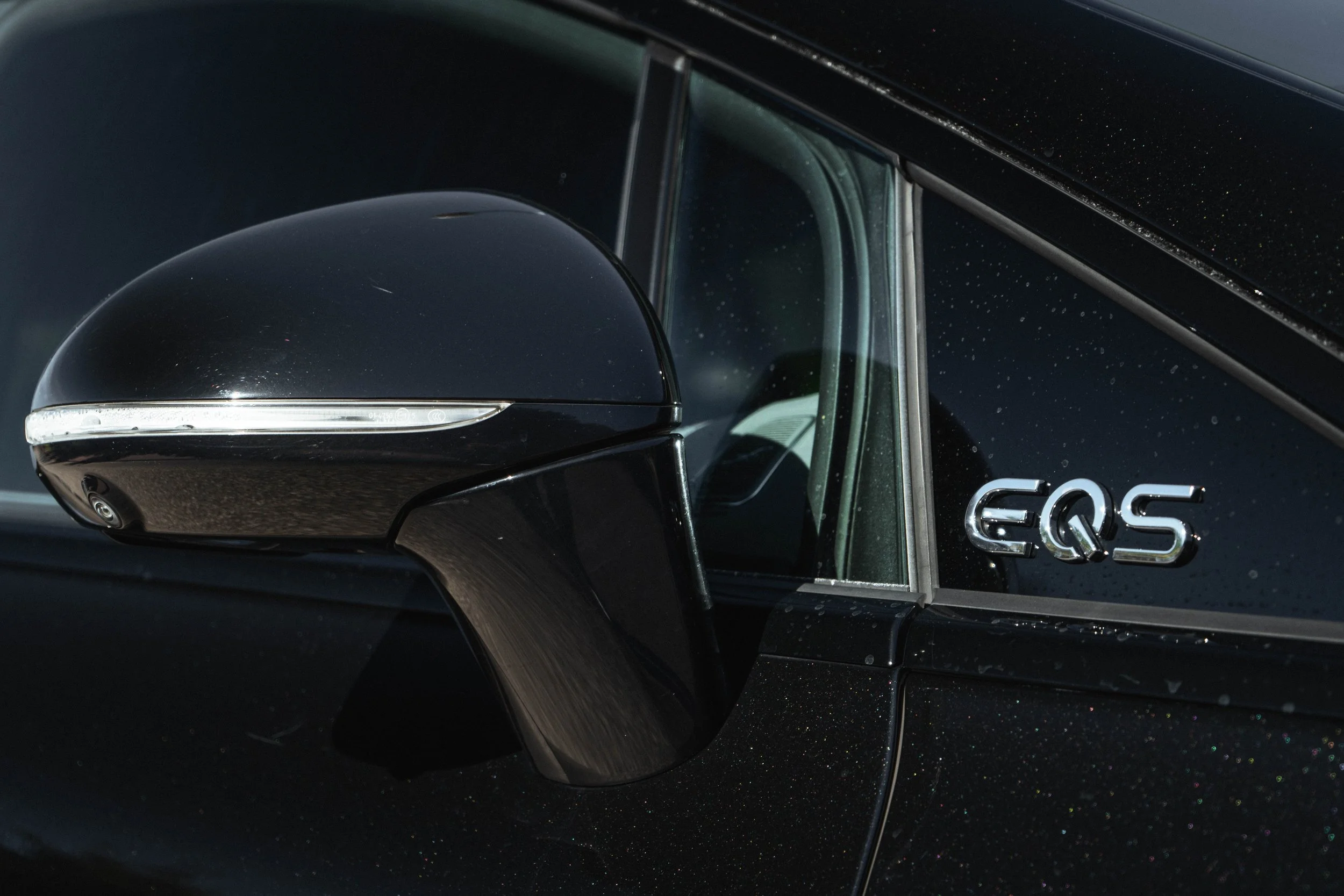 Espelho retrovisor de carro preto com acabamento em prata e a marca EQS no lado da porta ao lado do vidro aplicado na janela.
