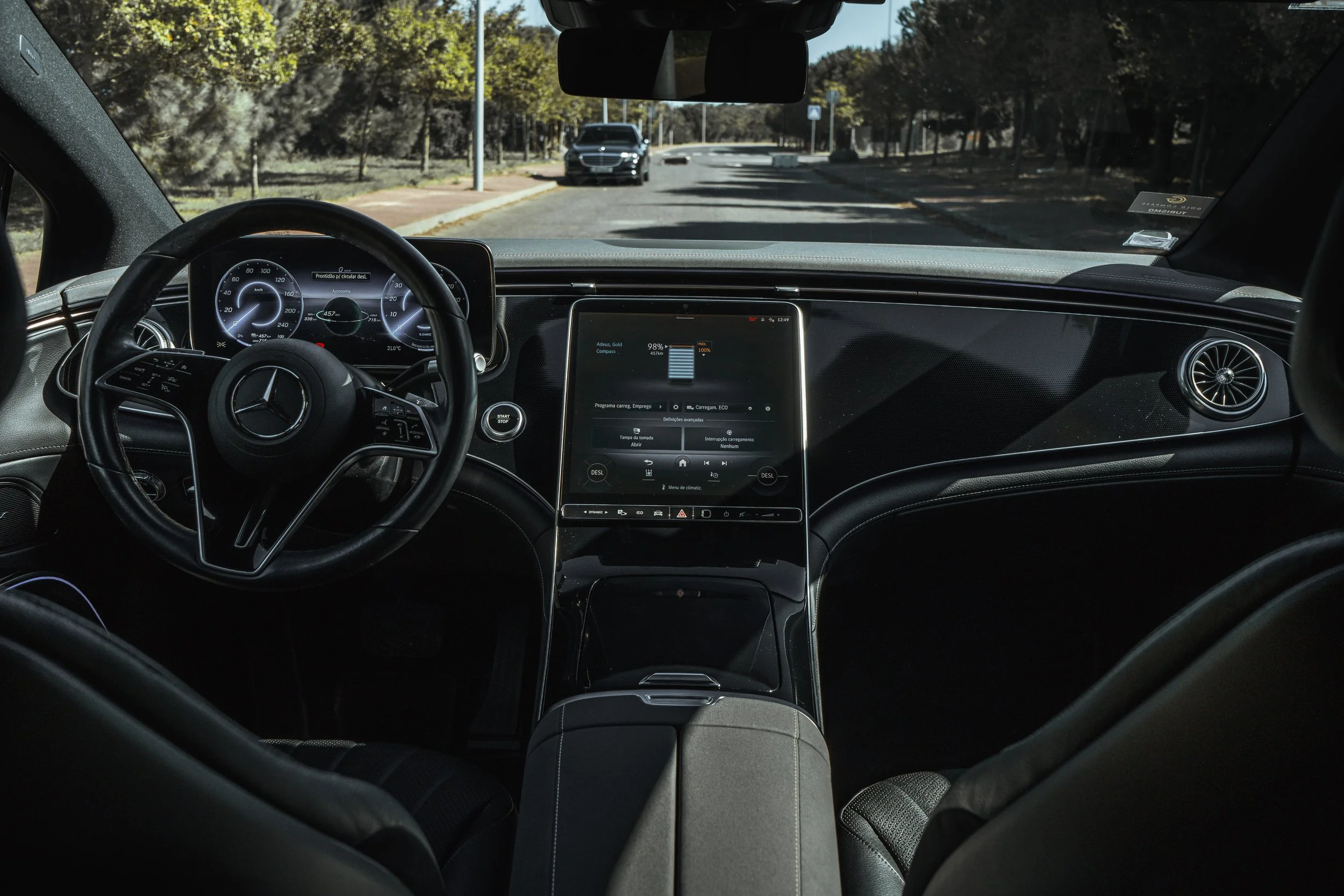 Interior de um carro de luxo Mercedes-Benz, mostrando o painel digital, volante, tela de comando central e assentos de couro, com uma rua arborizada visível pela frente.