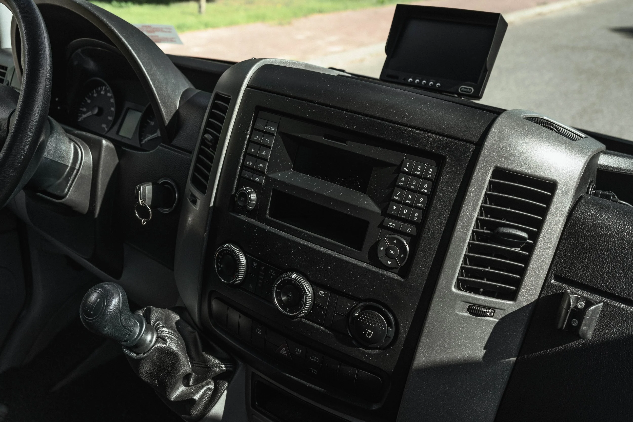 Interior de um caminhão com painel de controle, volante, câmbio manual e um monitor na parte superior.