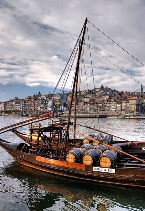 Embarcação de madeira com barris de vinho no rio em frente a uma cidade colorida, sob céu parcialmente nublado.