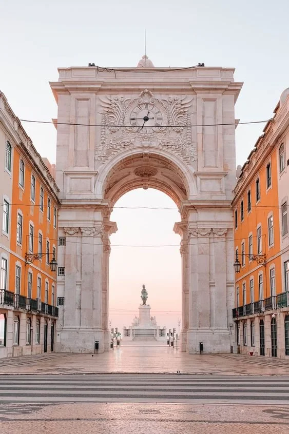Arco do Triunfo de Lisboa com uma praça em frente e uma estátua ao fundo, ao amanhecer ou entardecer, cercado por edifícios de cor laranja.