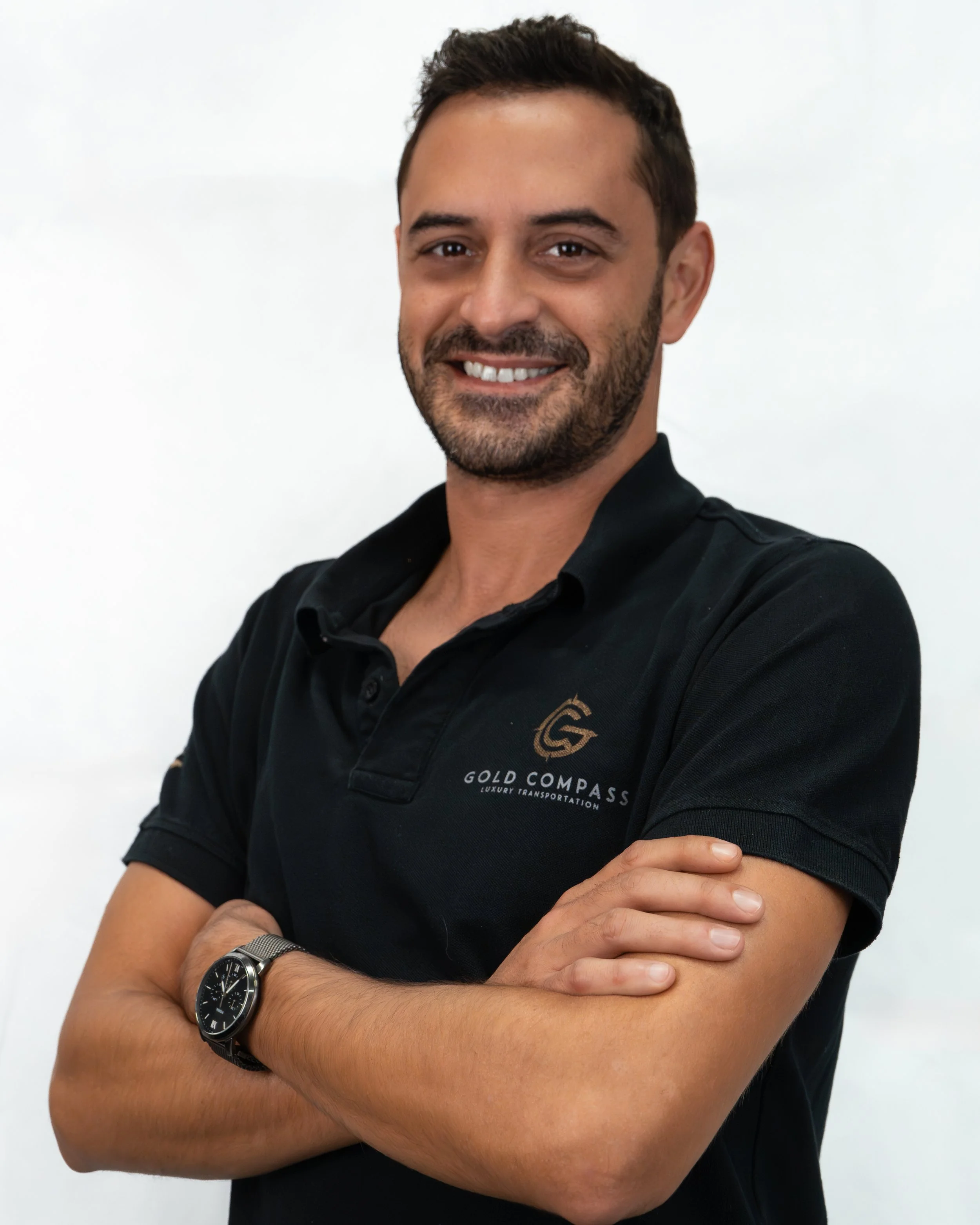 Homem sorridente com barba, usando camiseta preta com logo da Gold Compass, braços cruzados, usando relógio.