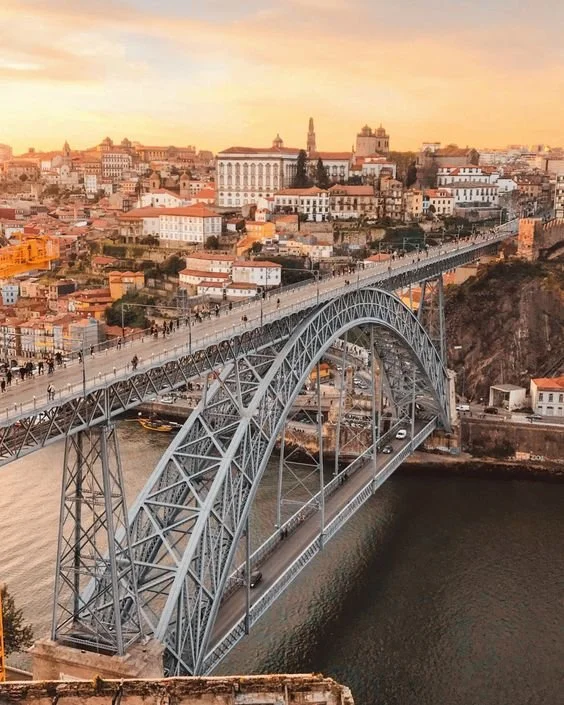 Ponte de arco de metal sobre um rio, com cidade ao fundo ao entardecer.