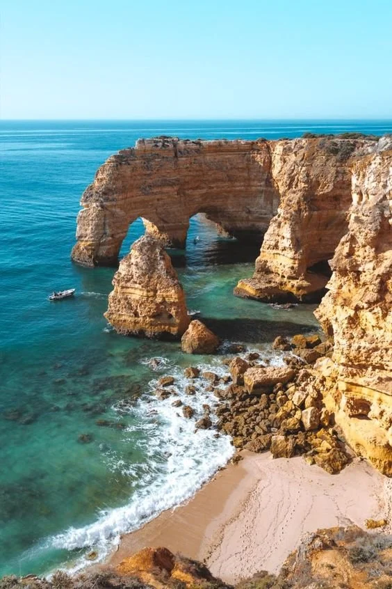 Formações rochosas com arcos na costa, água cristalina, pequena praia de areia dourada ao fundo, céu limpo.