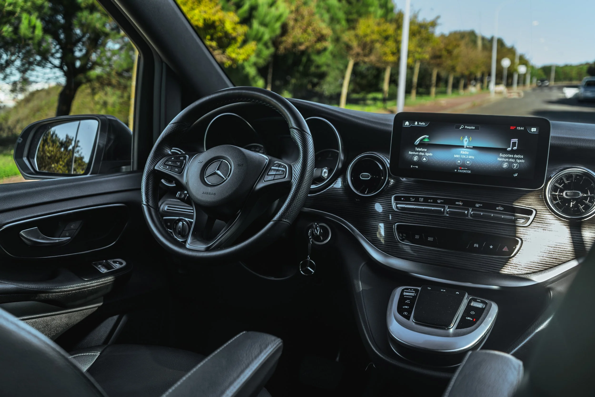 Interior de um carro Mercedes-Benz com painel digital e volante, visto de dentro na direção, em uma estrada cercada de árvores.