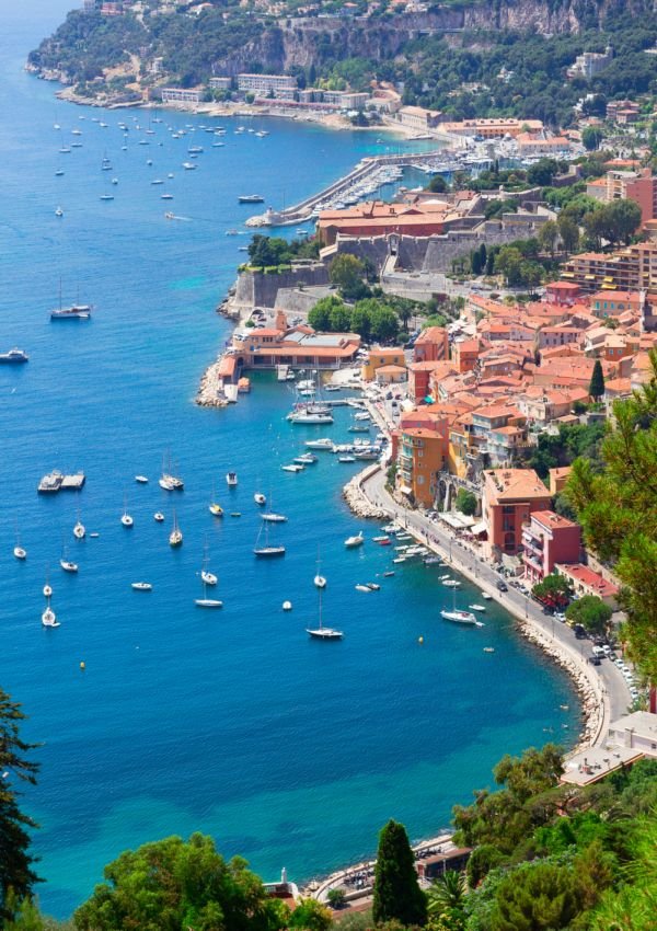 Cais com vários barcos e iates, casas coloridas pelo relevo, muro de fortificação antiga na encosta e vegetação verde ao redor.