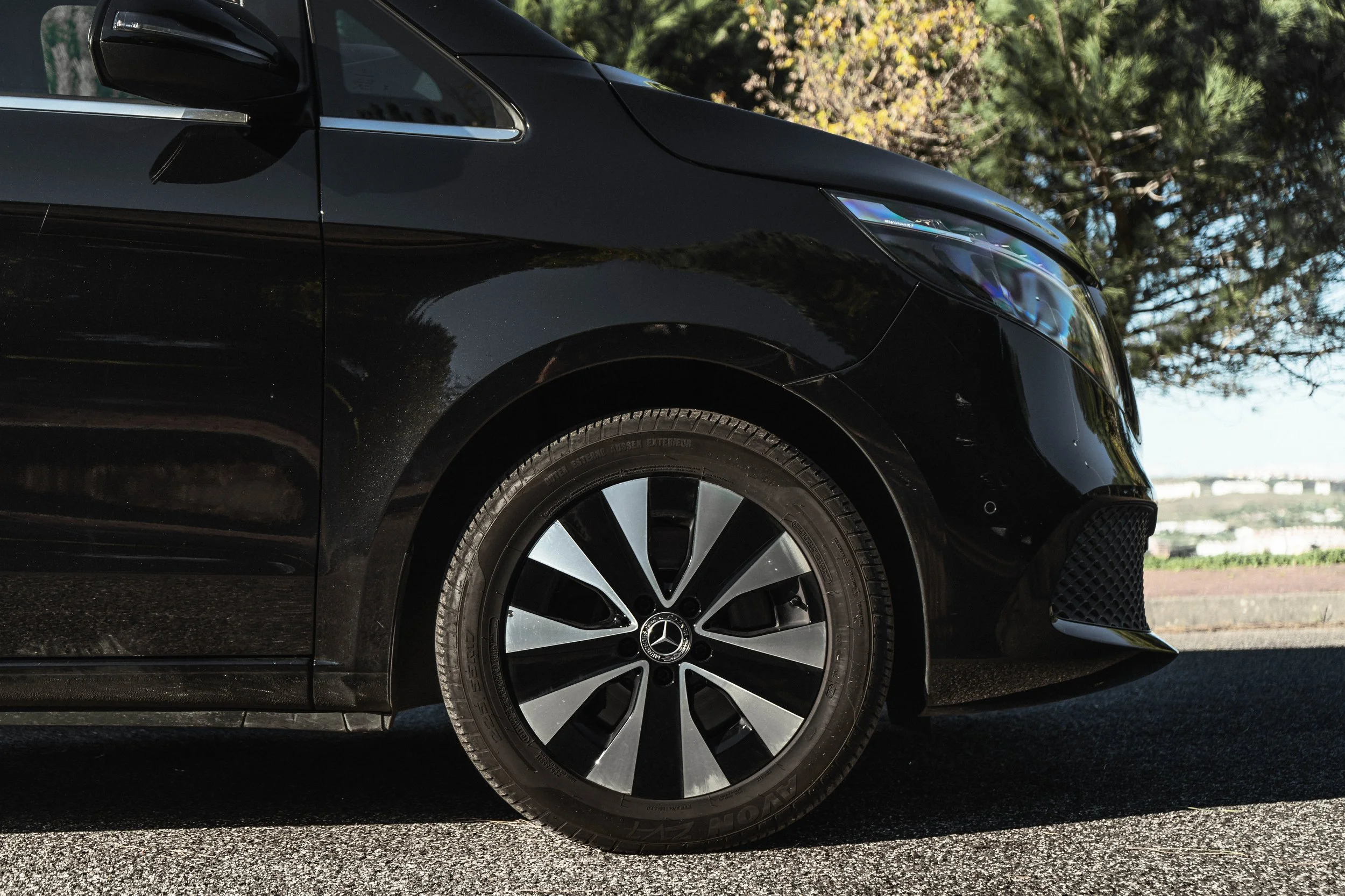 Carro preto, de frente, com roda de desenho moderno e logotipo da Mercedes-Benz, estacionado na rua com árvores ao fundo.