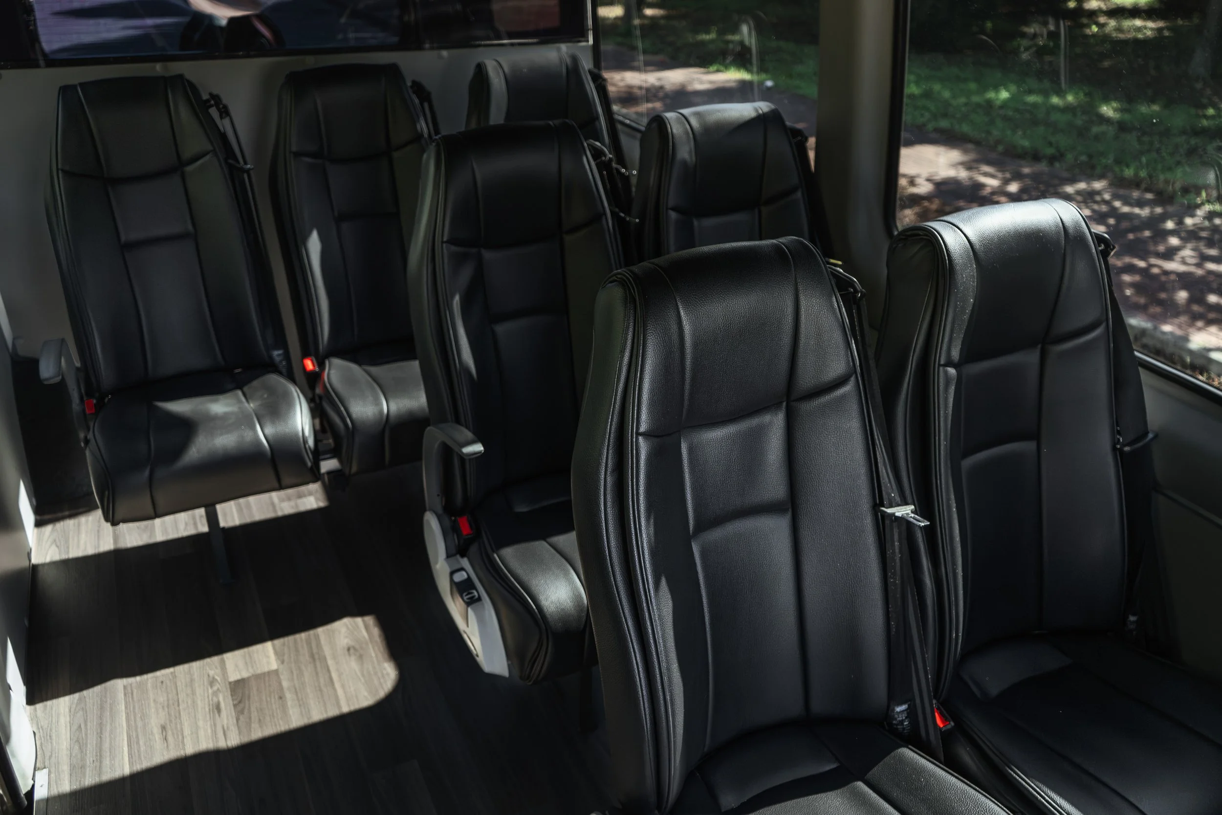 Interior de um ônibus com assentos de couro preto, alguns com braço articulado, próximo a janelas grandes com vista para o exterior com árvores em movimento.