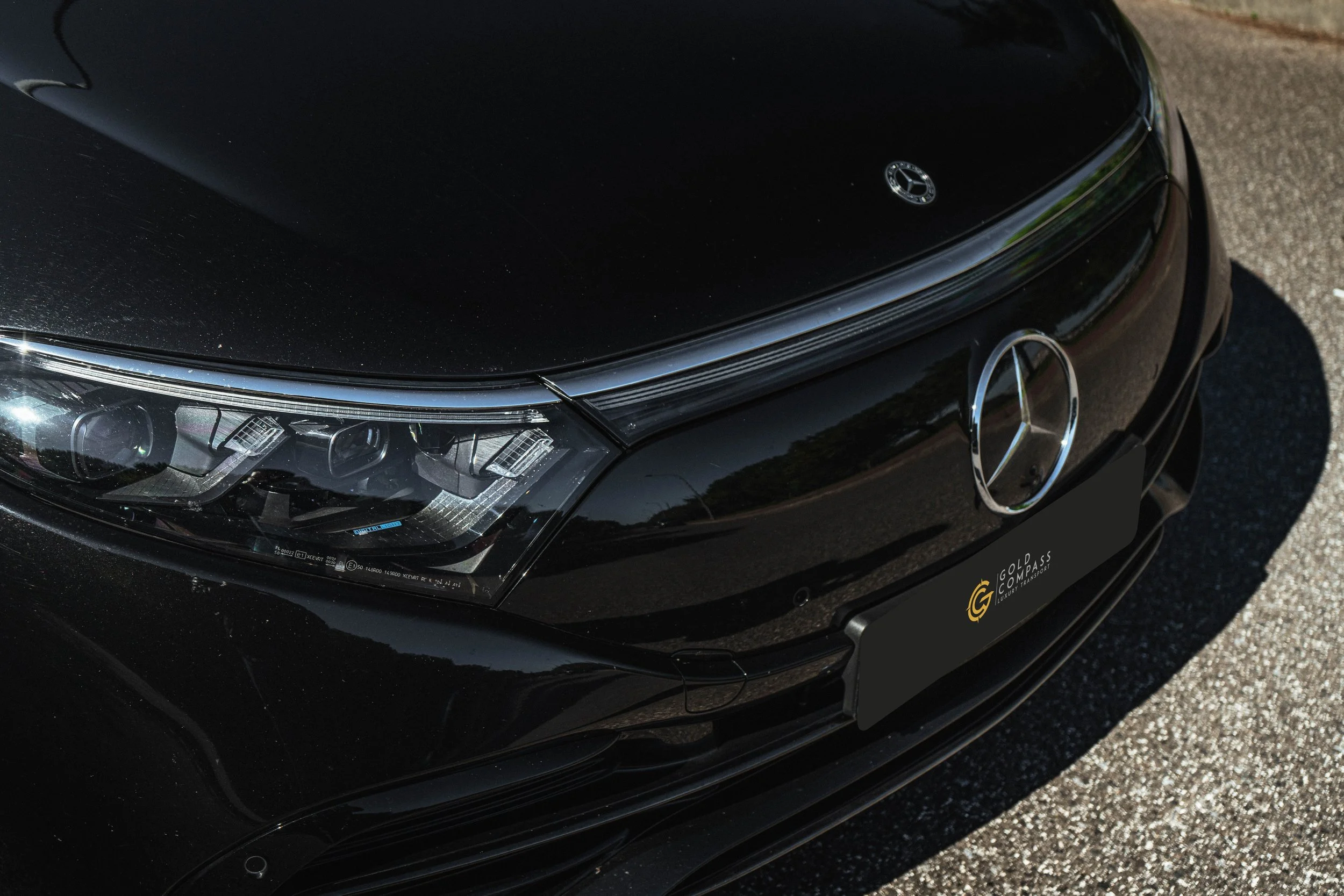 Fachada frontal de um carro preto, Mercedes-Benz, com destaque para o farol e o logotipo na grade dianteira, em piso de asfalto.