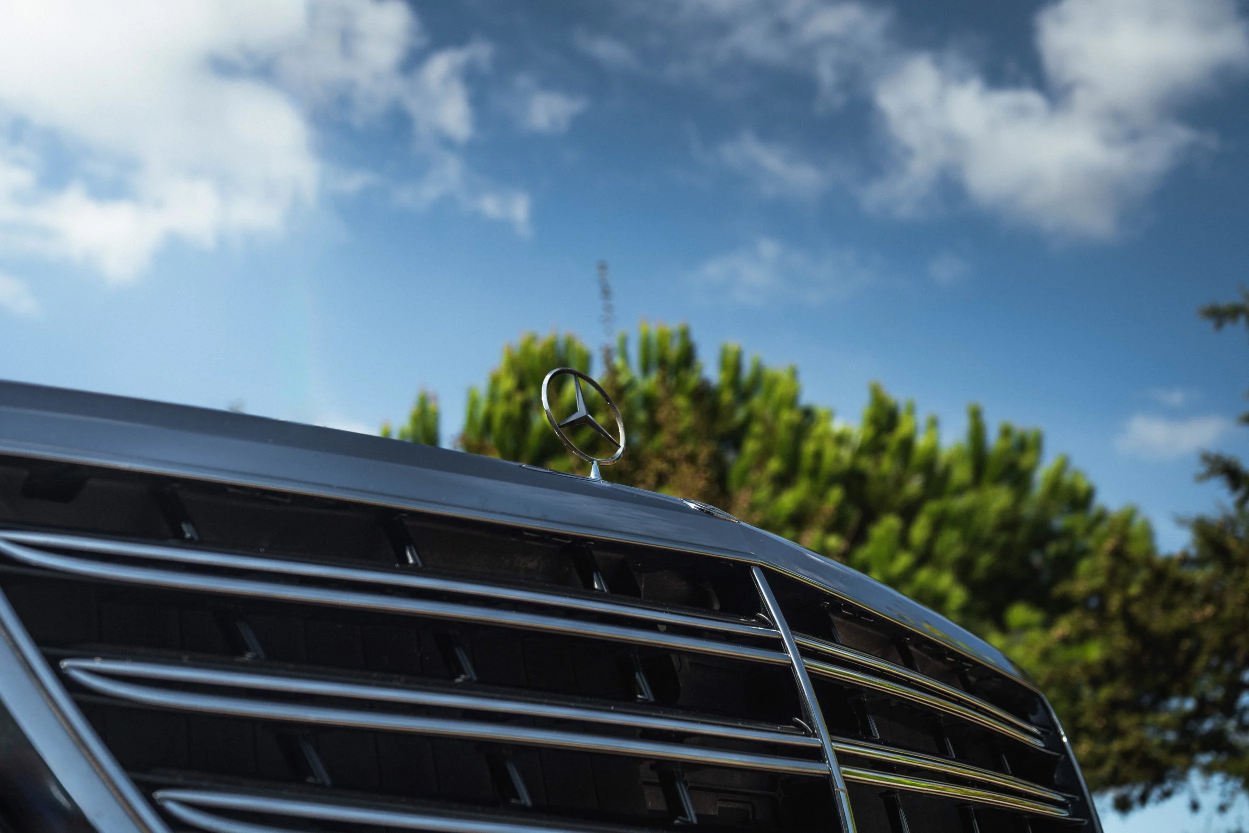 Fachada de carro Mercedes-Benz com logotipo na dianteira, sob céu azul com nuvens e árvores ao fundo.