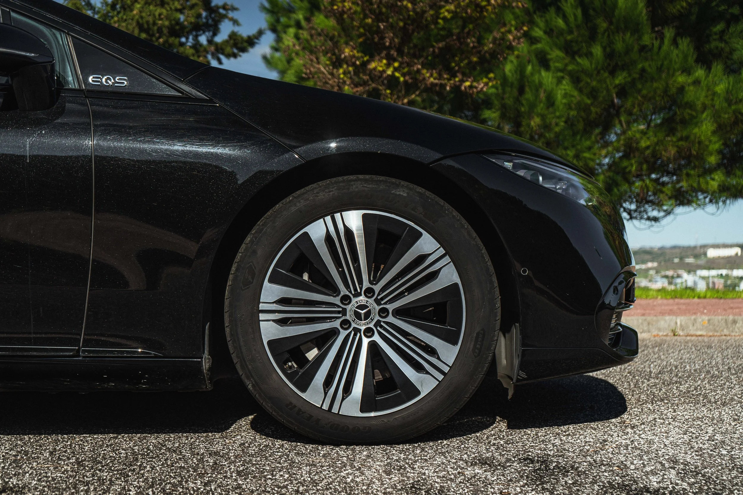 Parte frontal de um carro preto Mercedes-Benz, mostrando a roda, o farol e o lado da porta, estacionado na rua com árvores ao fundo.