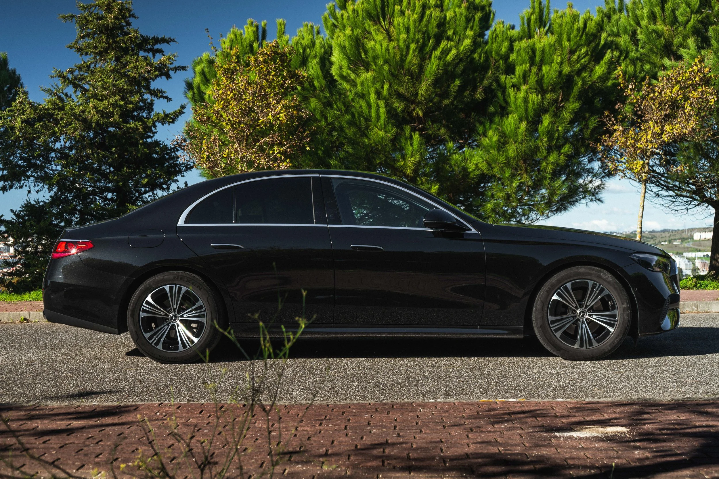 Carro preto sedã estacionado na rua com árvores verdes ao fundo em dia ensolarado.