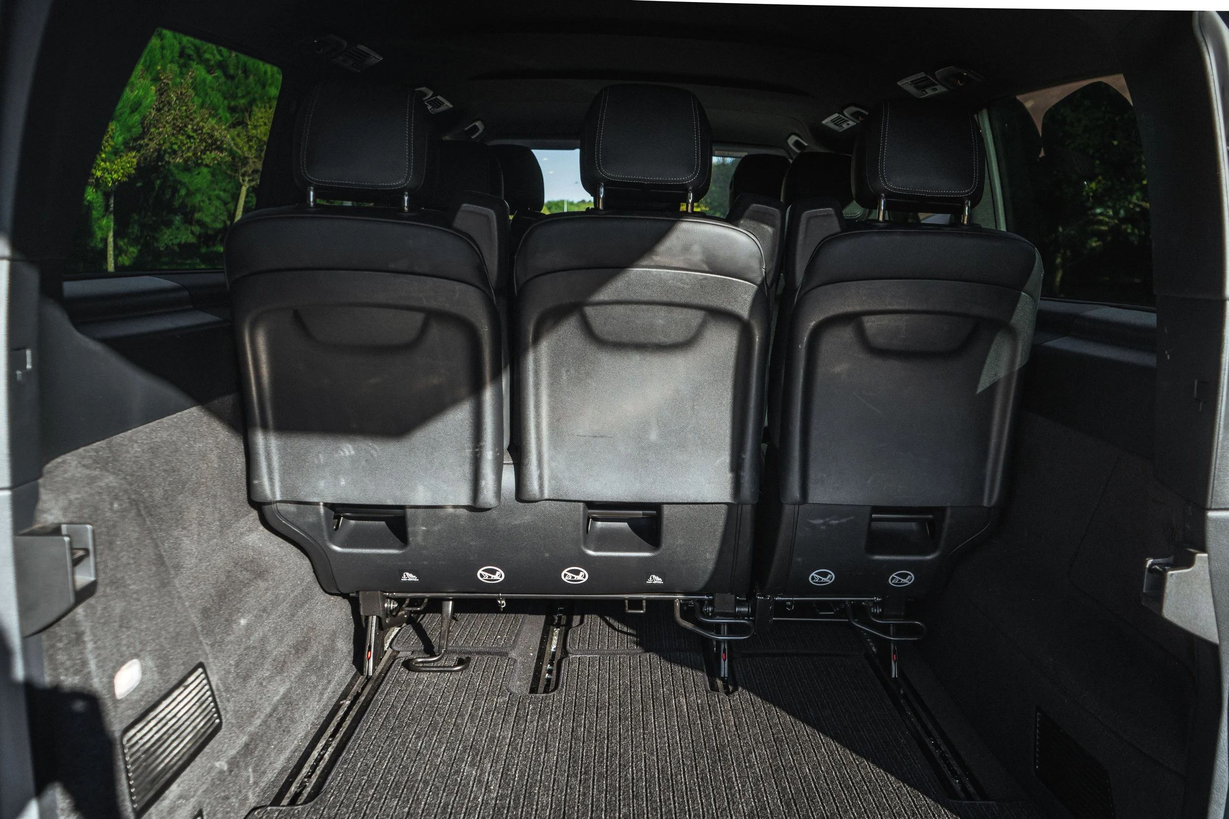 Interior de um veículo com assentos pretos e espaço de carga na parte traseira, com janelas laterais mostrando árvores ao redor.