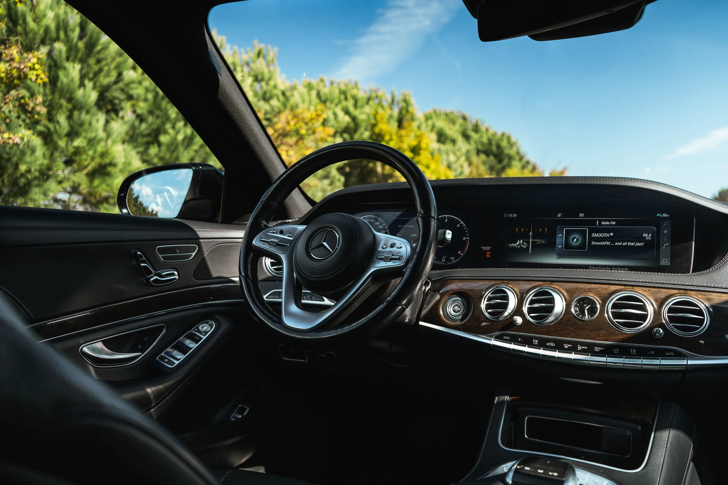 Interior do carro Mercedes-Benz mostrando painel, volante e tela de entretenimento, com visibilidade do exterior de árvores verdes e céu azul ao fundo.