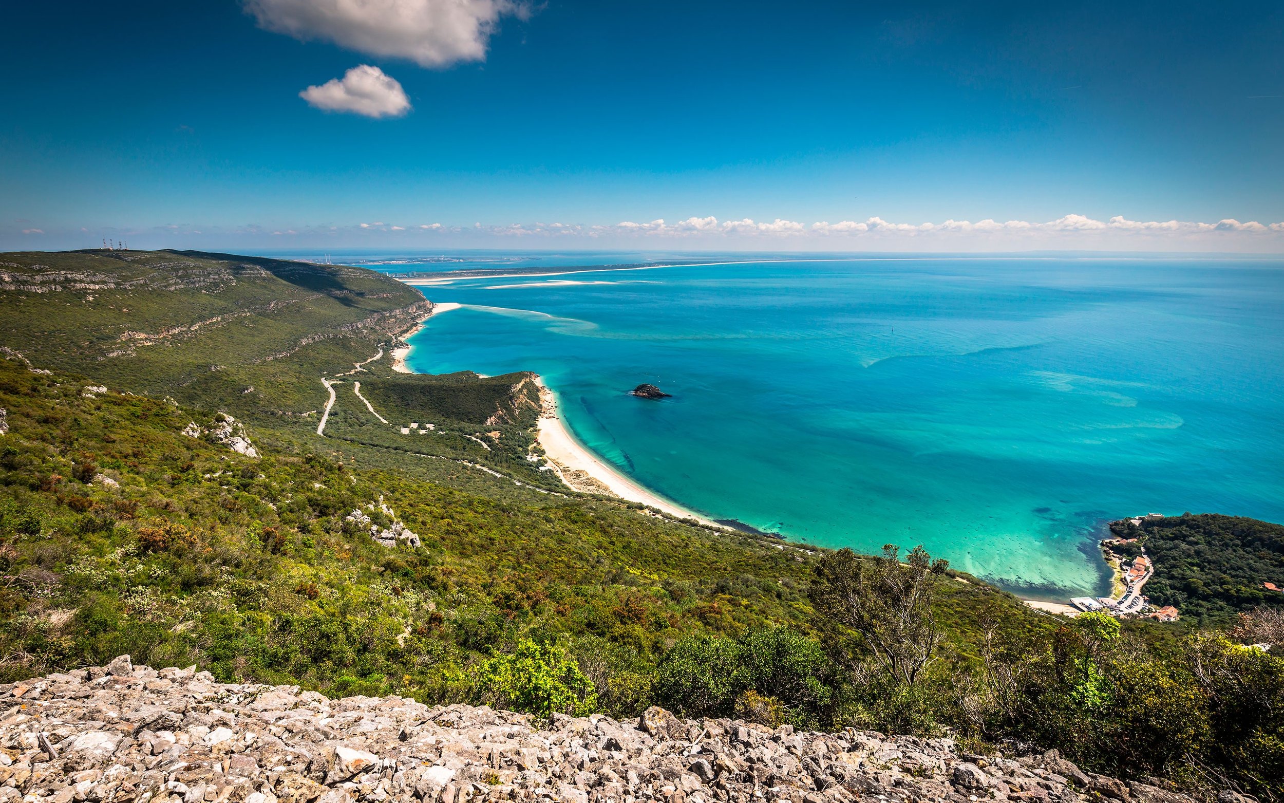 Vista de uma costa com mar azul claro, praias de areia branca, vegetação verde e pequenas construções à beira-mar, sob céu azul com algumas nuvens.