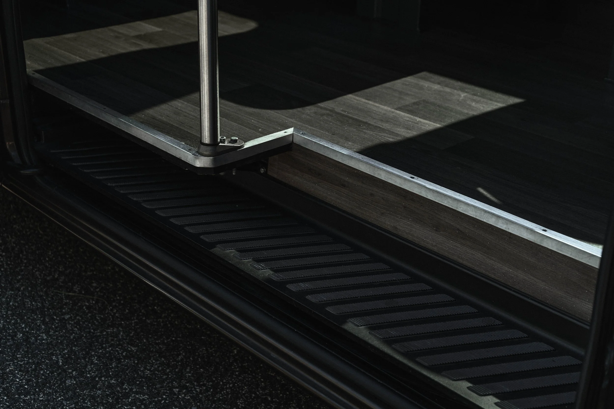 Detalhe da escada de um ônibus, com piso de madeira e degraus de metal, ao lado de uma plataforma de entrada.