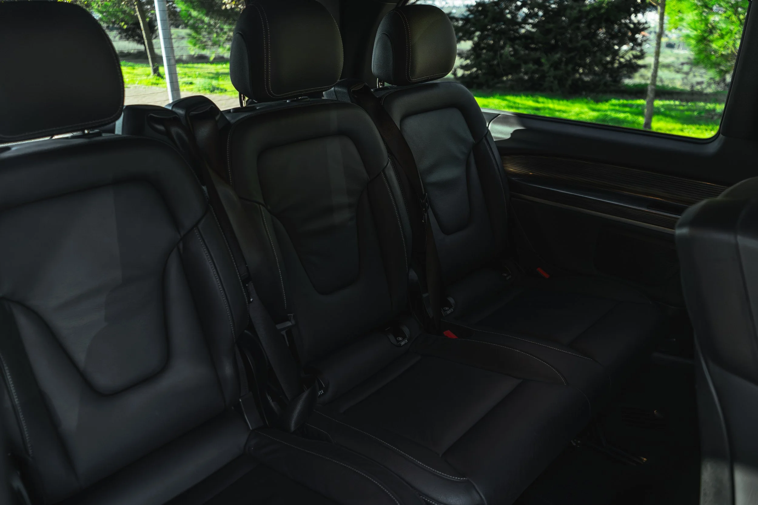 Assento de carro com três lugares, revestido em couro preto, com cabeçares ajustáveis e cintos de segurança. Janela ao lado revelando ambiente externo com árvores e grama verde.