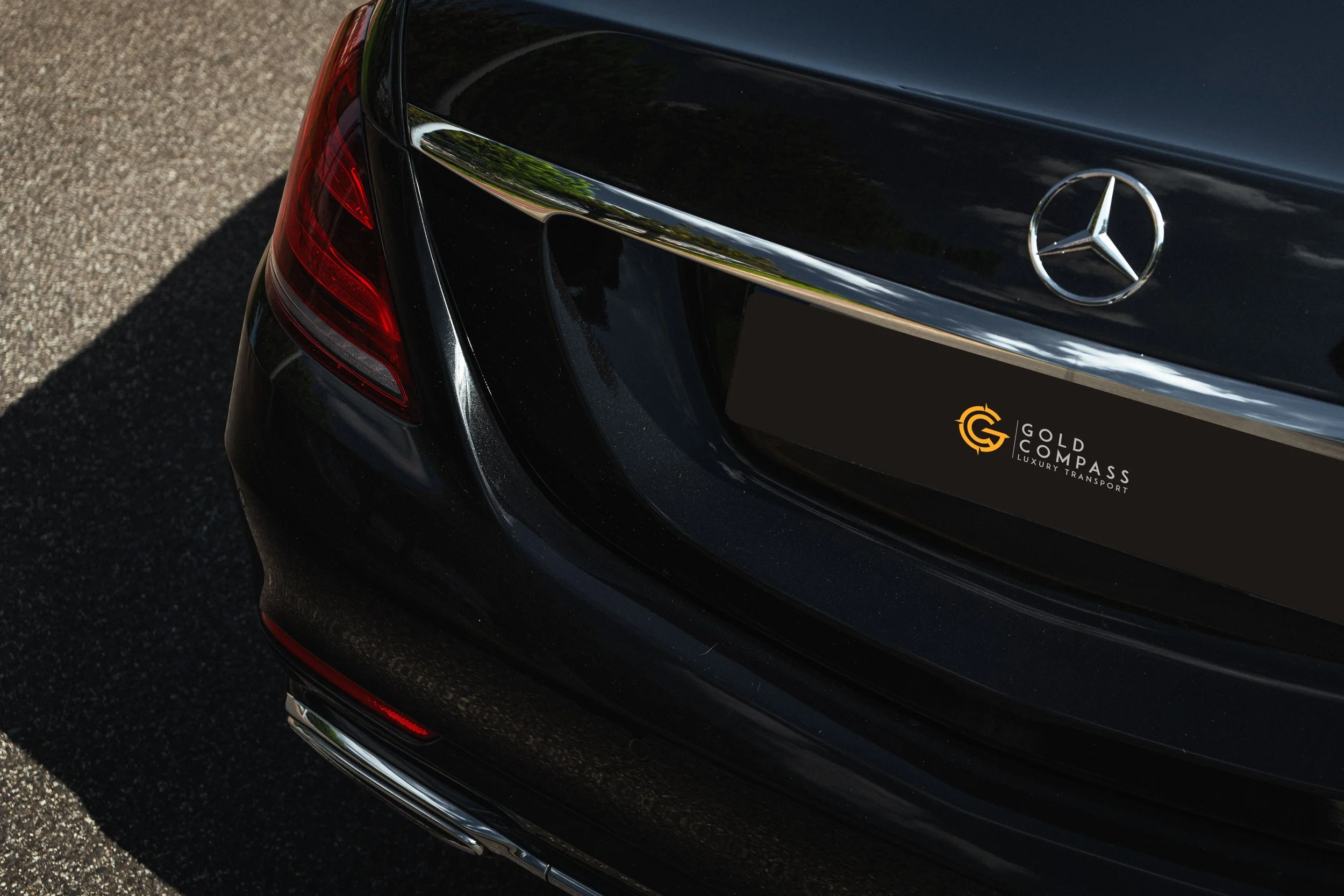 Parte da traseira de um carro preto, com o logo da Mercedes-Benz. Há uma inscrição no porta-malas que diz Gold Compass Luxury Transport.