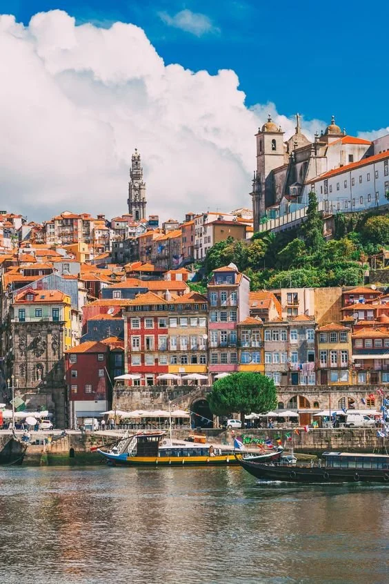 Paisagem urbana de cidade litorânea com edifícios coloridos, barcos no rio e uma catedral no topo da colina