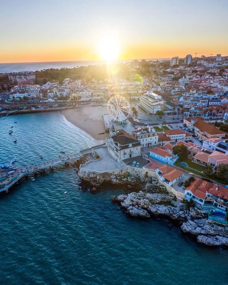 Vista aérea de uma cidade costeira ao pôr do sol, com roda gigante, mar, praia, prédios e casas coloridas.